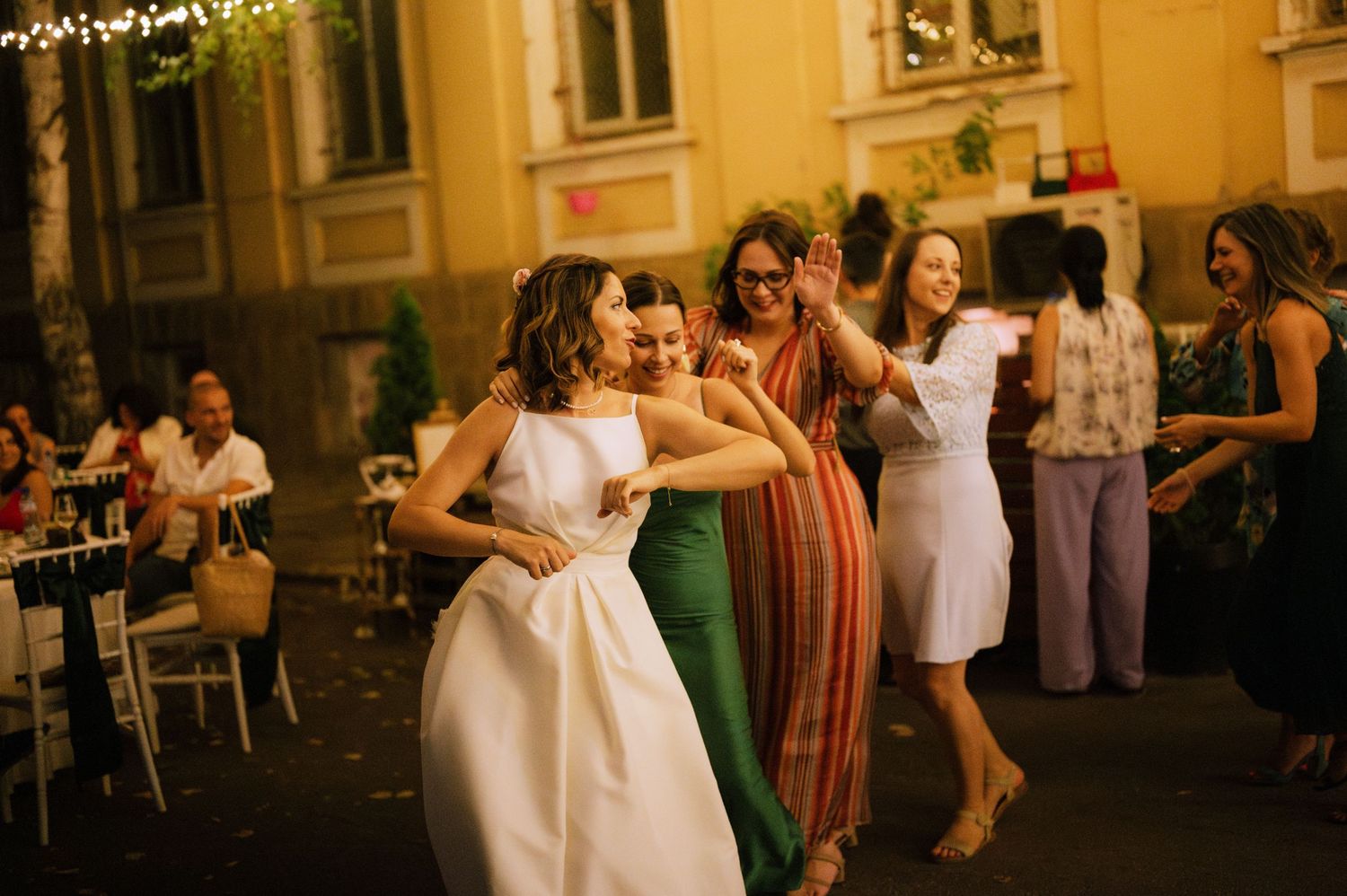 People dancing and celebrating at a golden-hour outdoor wedding reception with string lights overhead.