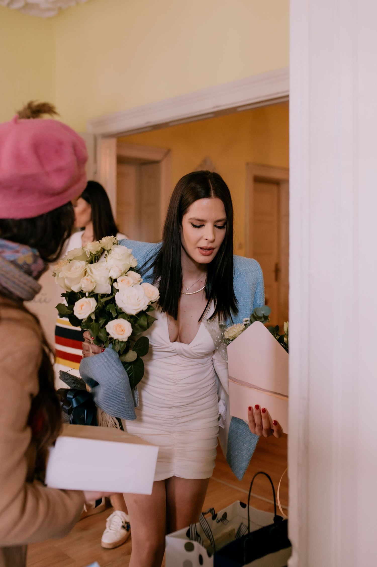 A woman in a white dress and denim jacket holds a bouquet of pink and white flowers in a hallway.