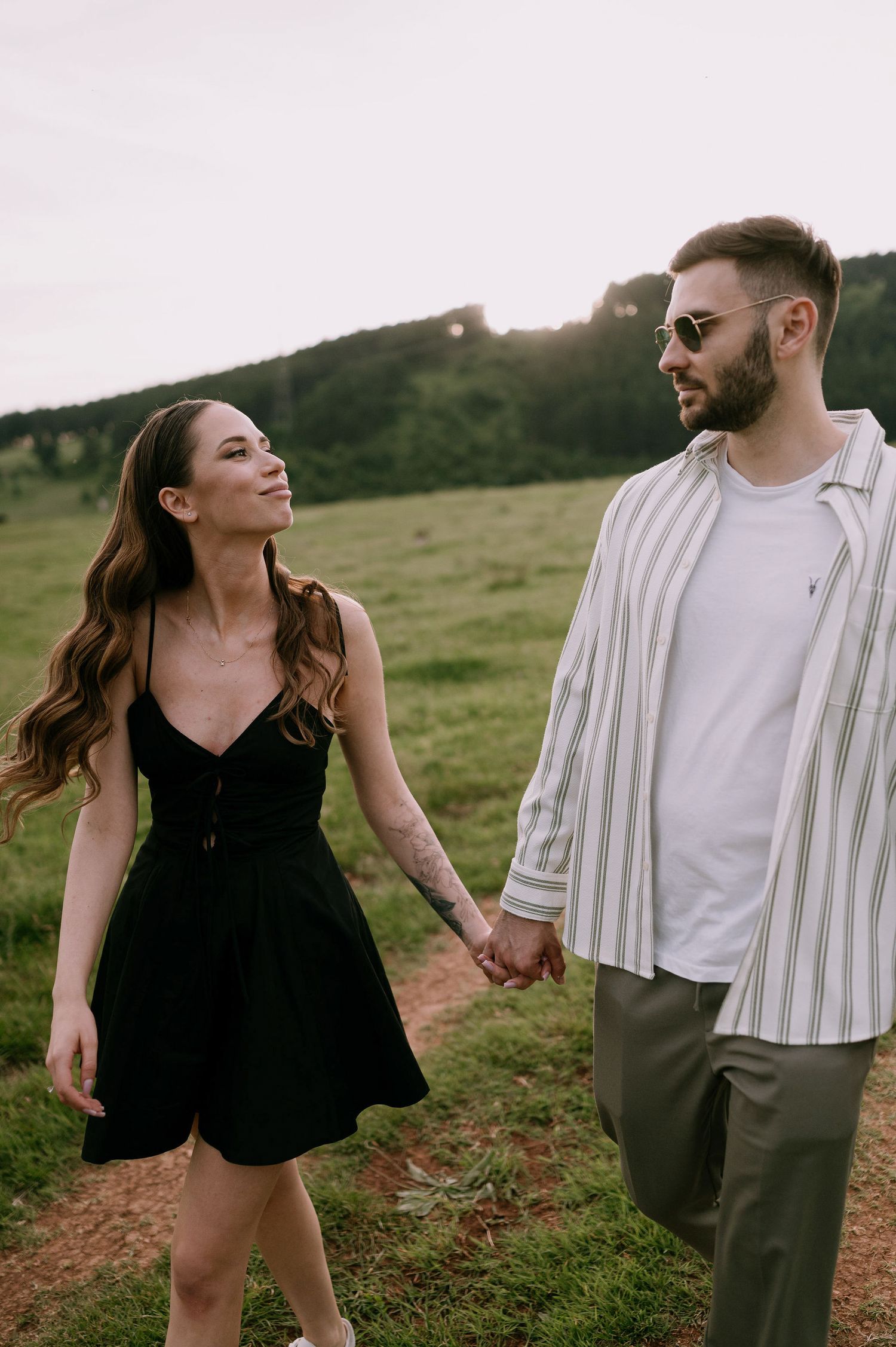 A couple in a black dress and striped shirt hold hands while walking through a grassy hillside at sunset.