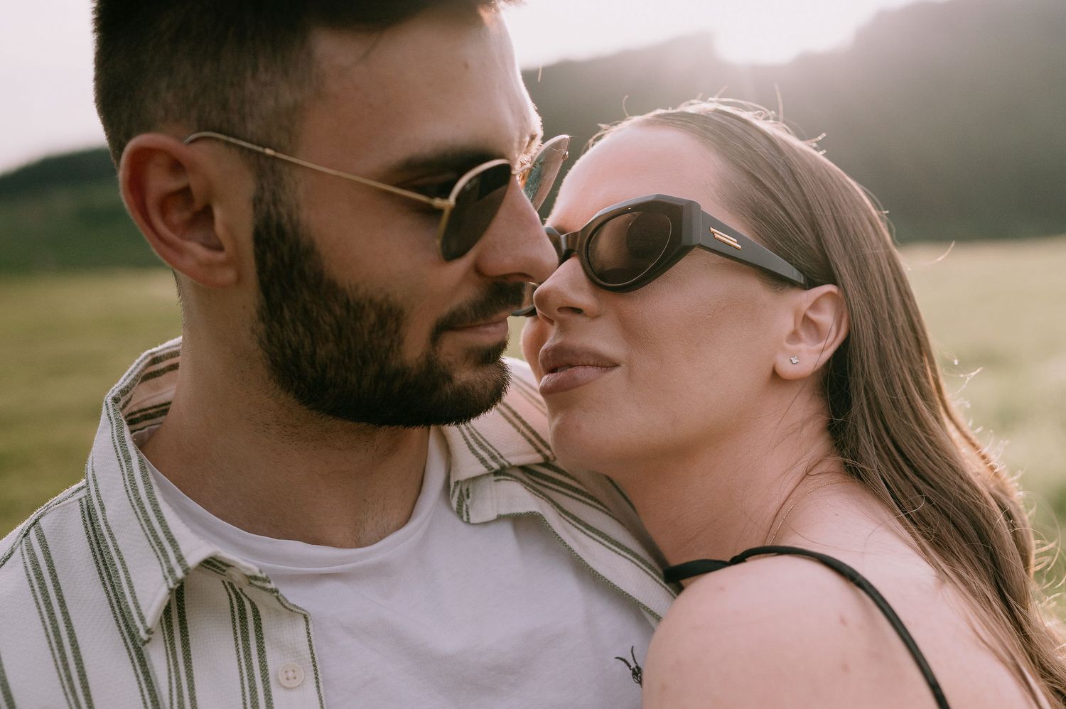 A romantic couple wearing sunglasses share an intimate moment together in warm sunlight outdoors.
