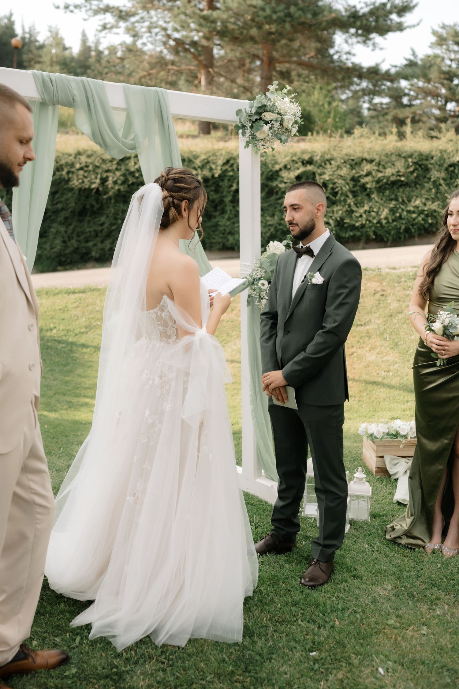 Outdoor wedding ceremony under white draped arch with couple and wedding party on grassy lawn surrounded by greenery.