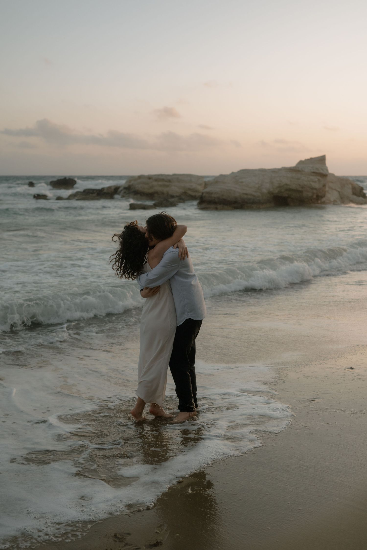 Couple embracing and kissing on a sandy beach at sunset, waves washing over their feet near rocky formations.