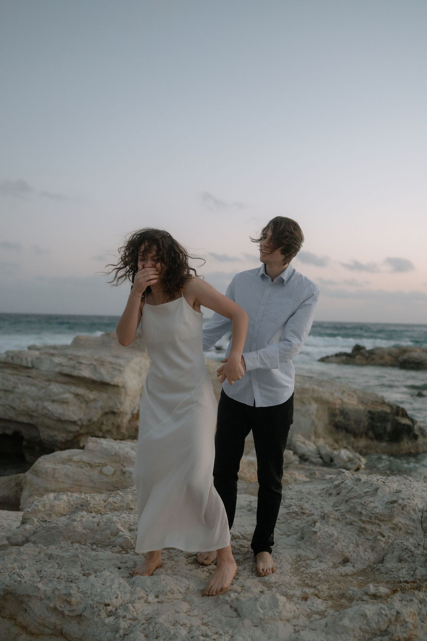Couple standing on rocky shore at dusk, woman in white slip dress, man in blue shirt, wind blowing their hair.
