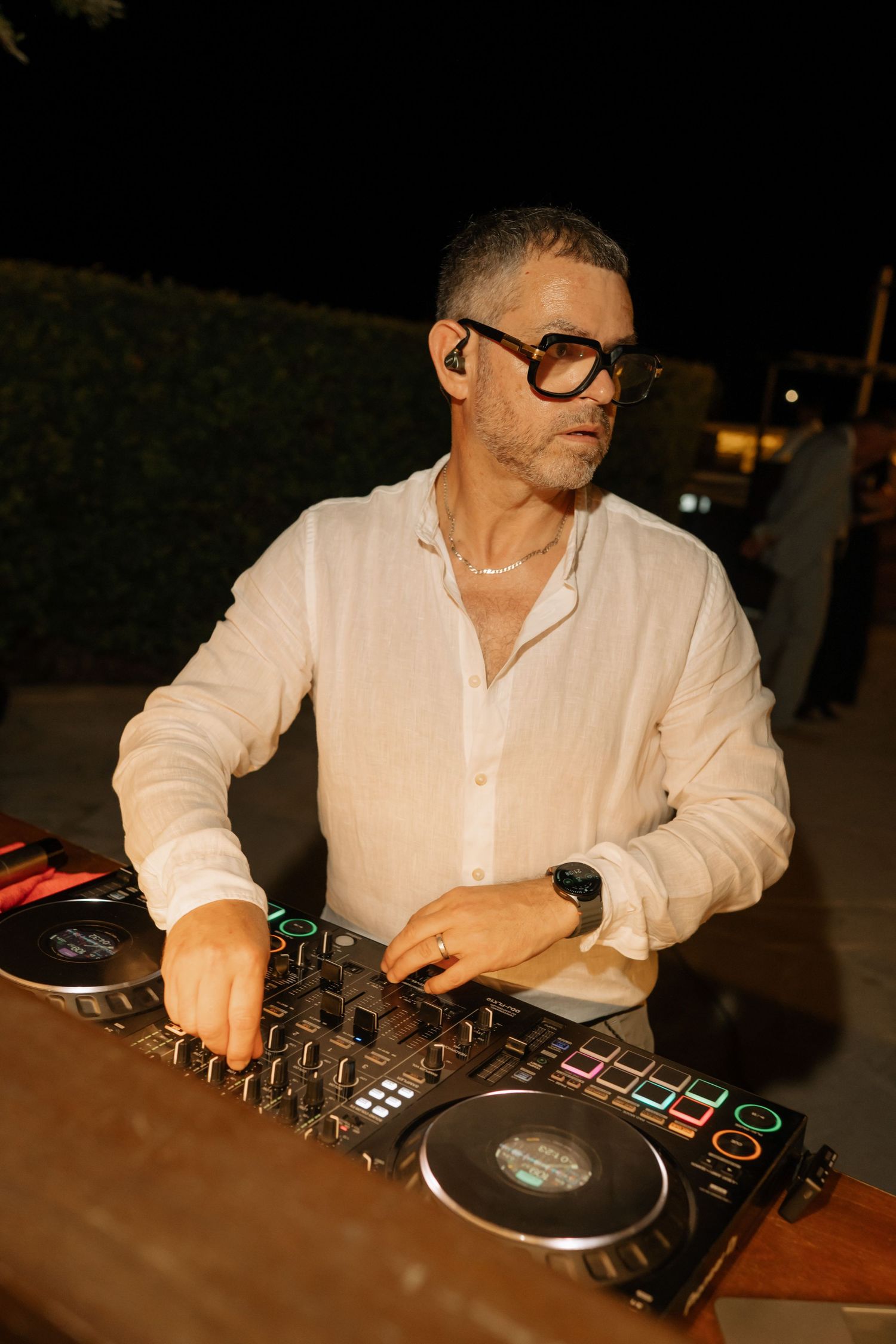 A DJ in white shirt and glasses mixing music on a controller at an outdoor nighttime event.