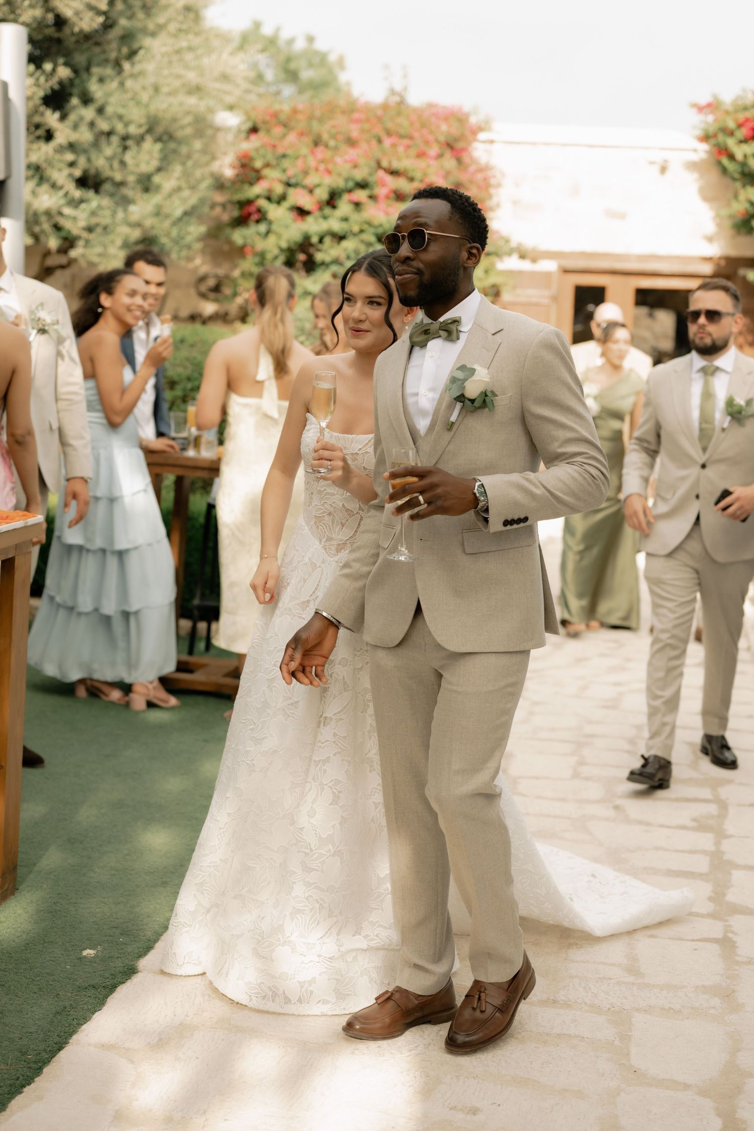 Bride and groom walking together at outdoor wedding reception, groom in beige suit with sunglasses, bride in white lace gown.