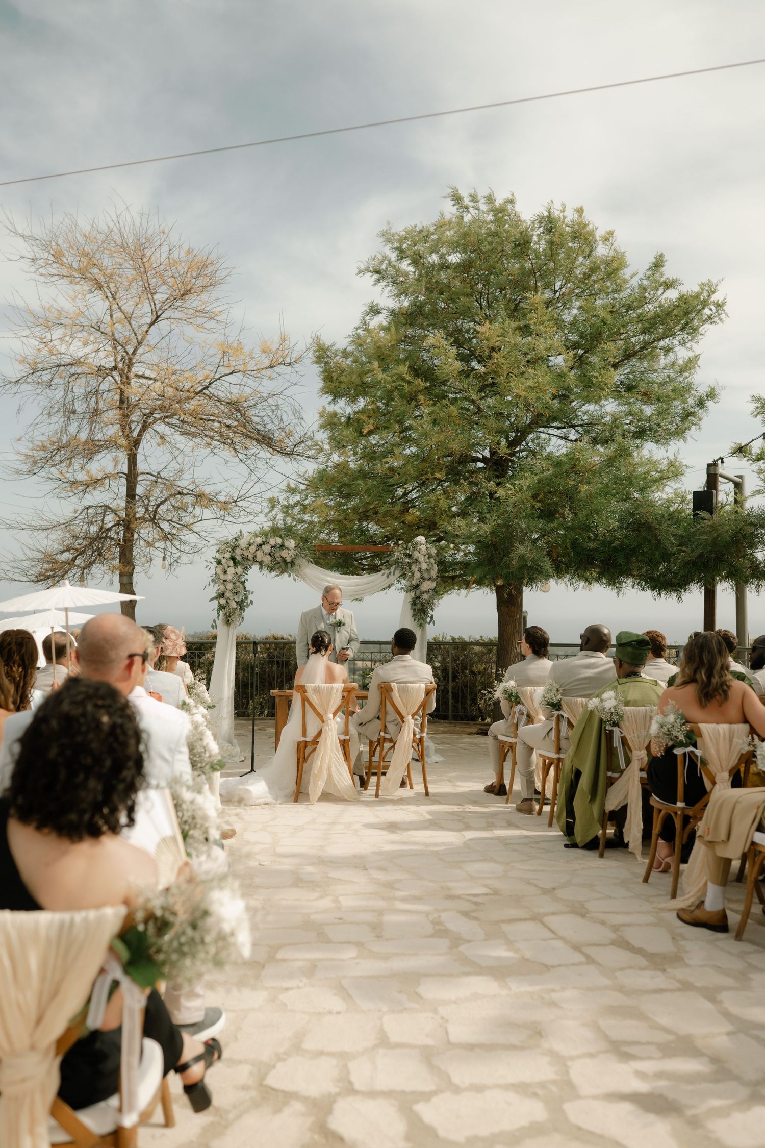 Outdoor wedding ceremony on stone patio with guests seated in wooden chairs facing couple at floral arch, surrounded by trees.
