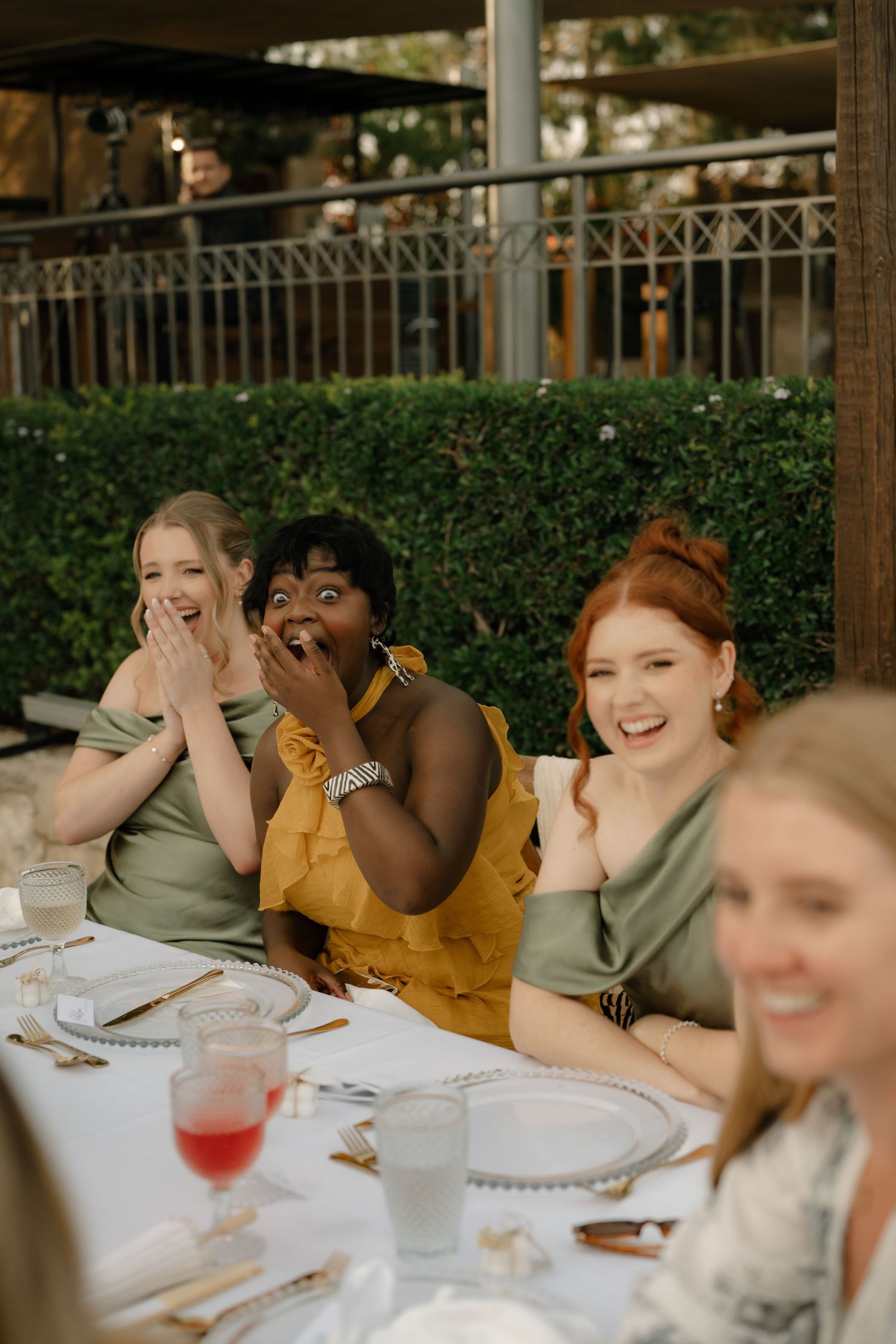 Three women laughing joyfully at an outdoor wedding reception dinner table, wearing sage green and yellow dresses.