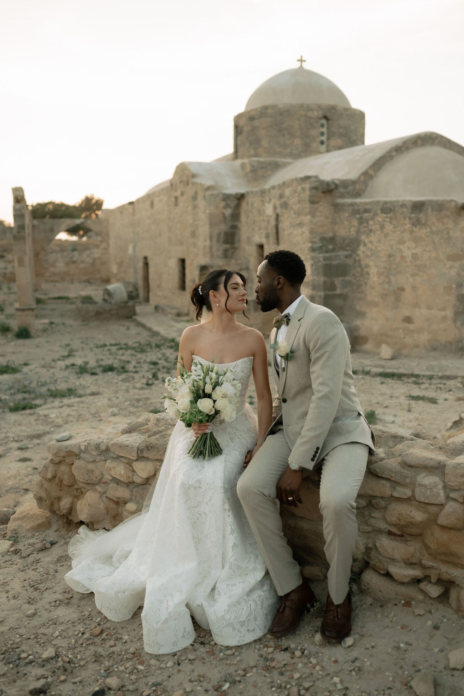 Wedding couple sharing a romantic kiss near ancient stone church ruins during golden hour sunset.