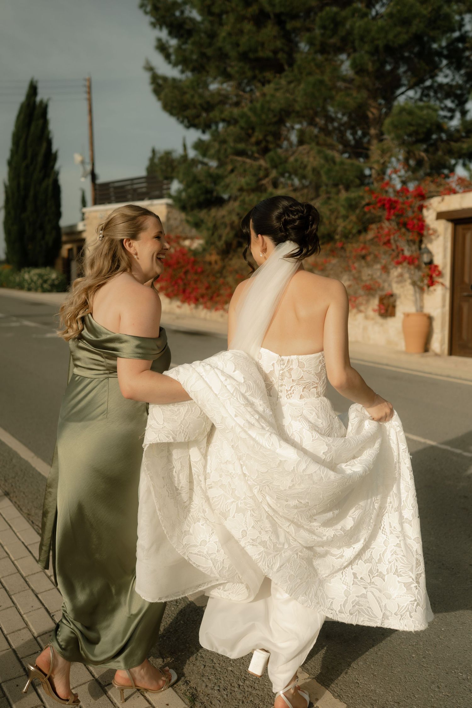 Bride in white lace gown walks with bridesmaid in green satin dress, holding her train on a sunny street.