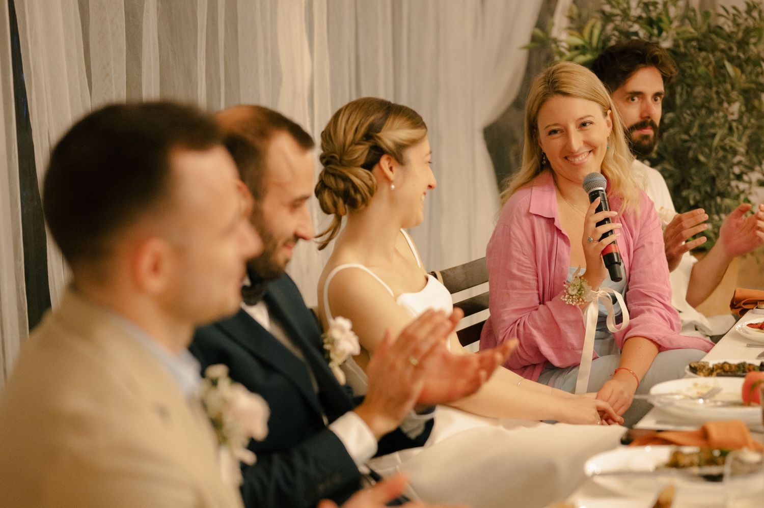 Wedding reception guests enjoying dinner and drinks at a long table during an evening celebration.
