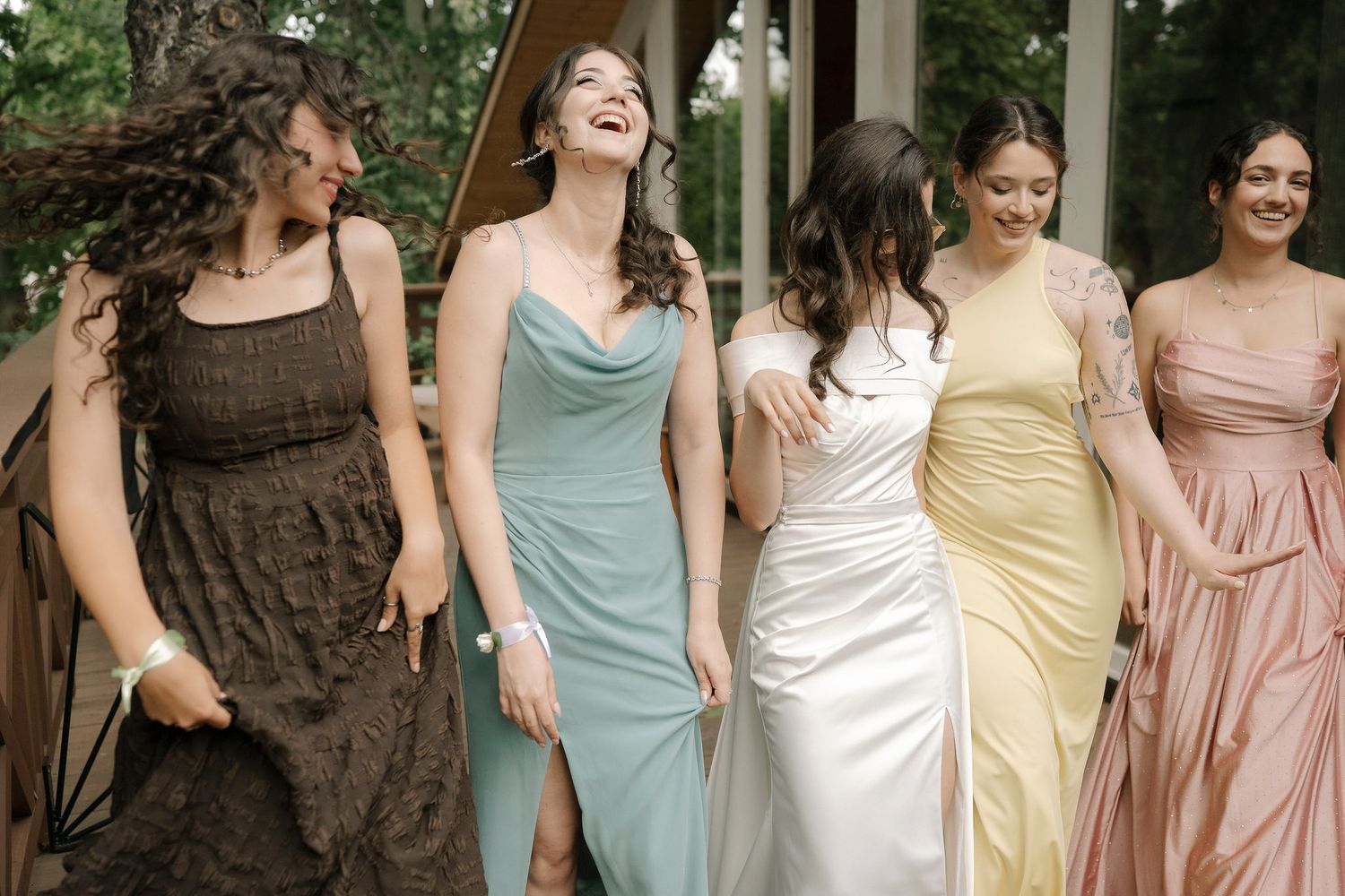A row of bridesmaids in pastel and brown dresses laugh together while walking outdoors.