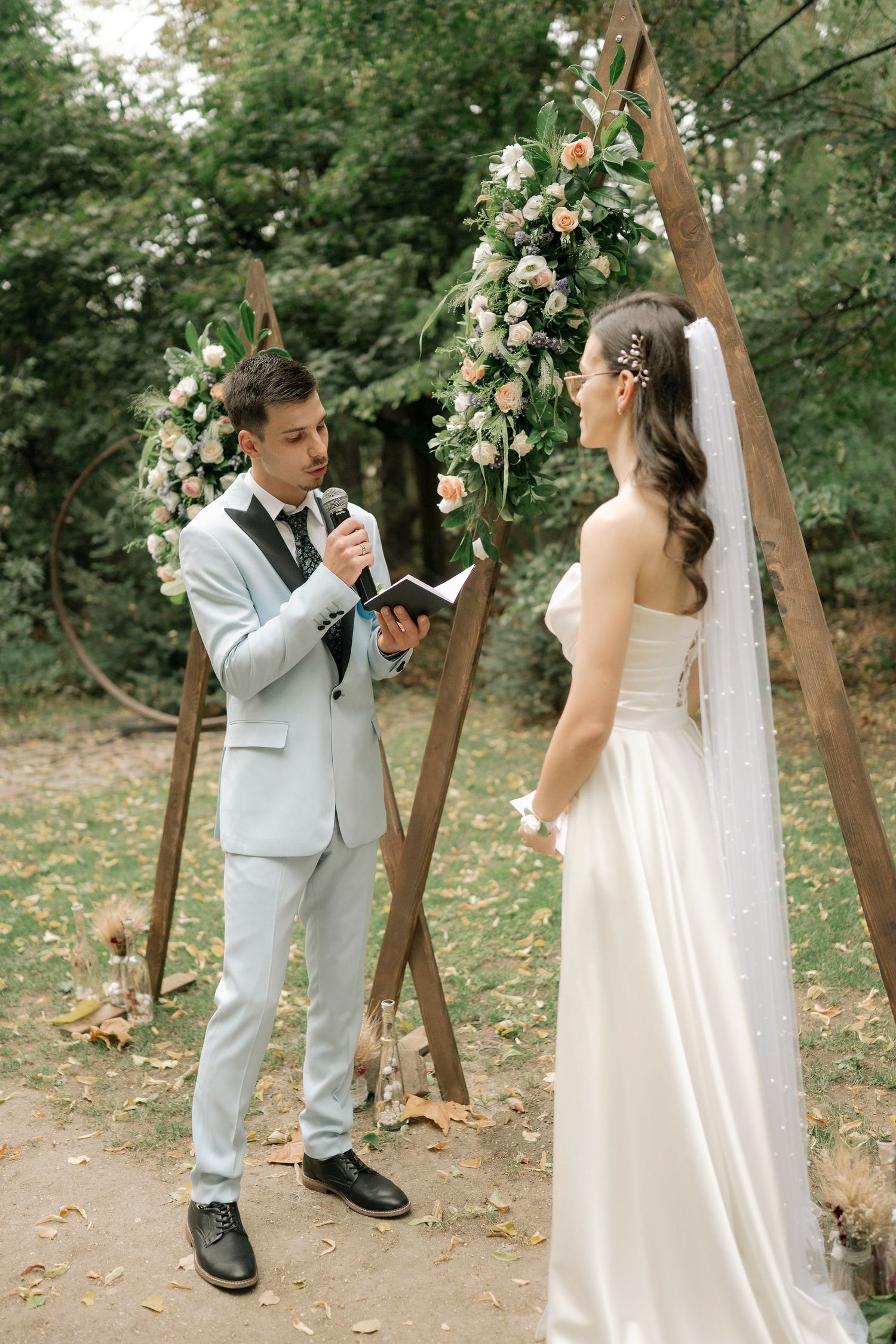 A wedding ceremony takes place beneath a rustic wooden arch adorned with white roses and greenery in a forest setting.