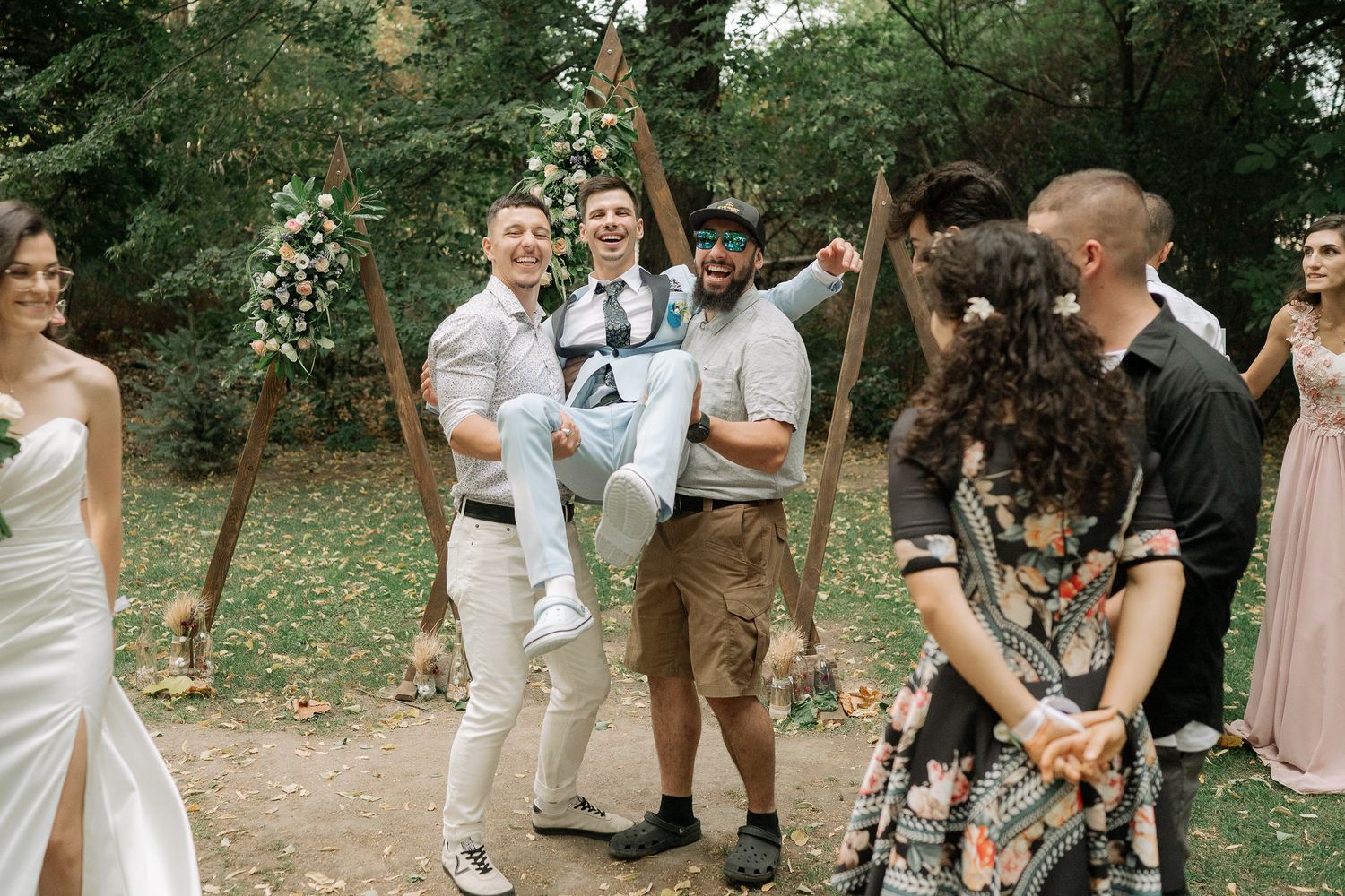 People gathered in a casual outdoor celebration wearing light colored clothing in a natural setting.