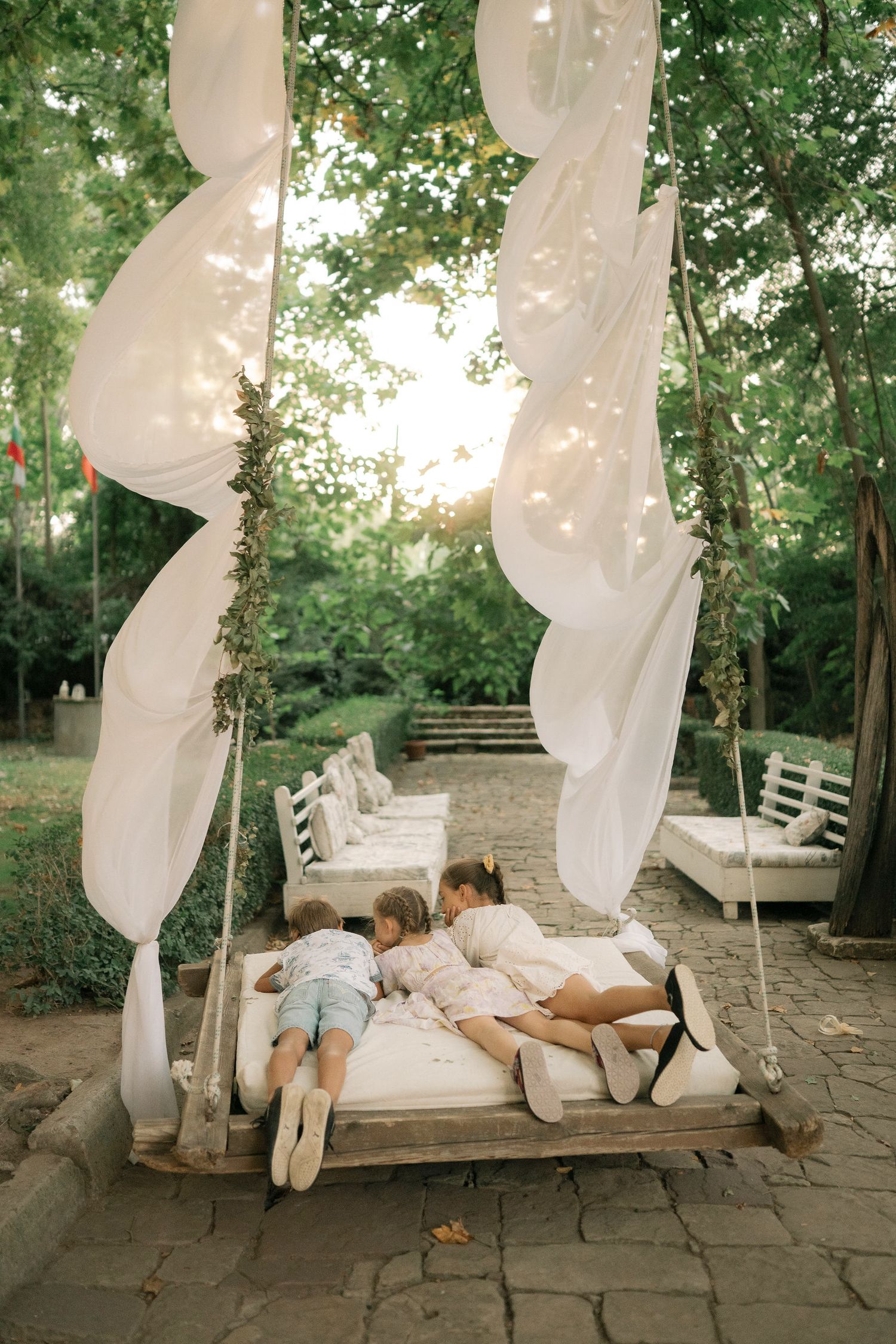 A dreamy outdoor swing bed with flowing white curtains and cushions in a lush garden setting during golden hour.
