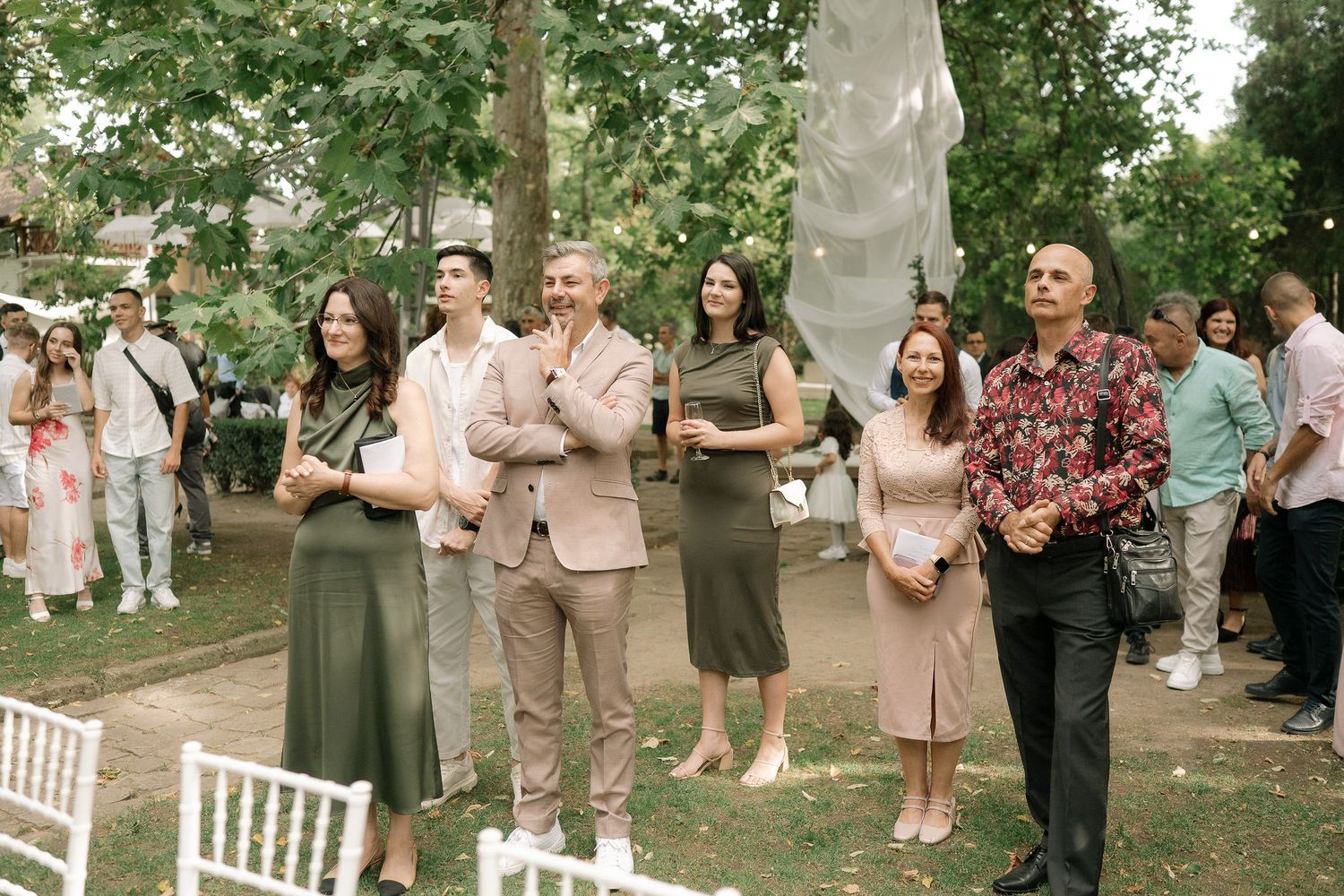 Wedding guests mingle and socialize during an outdoor garden ceremony with white chairs and lush greenery.