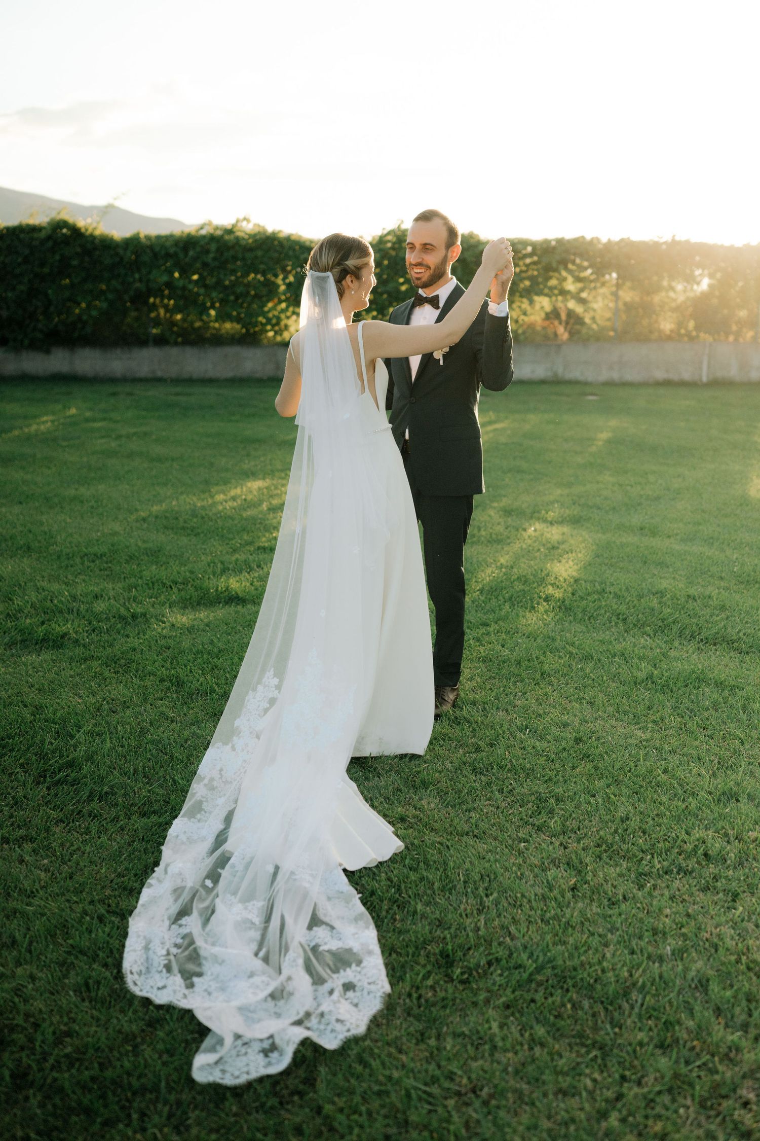 Newlywed couple walking together on grassy field at sunset with bride's lace dress train trailing behind.