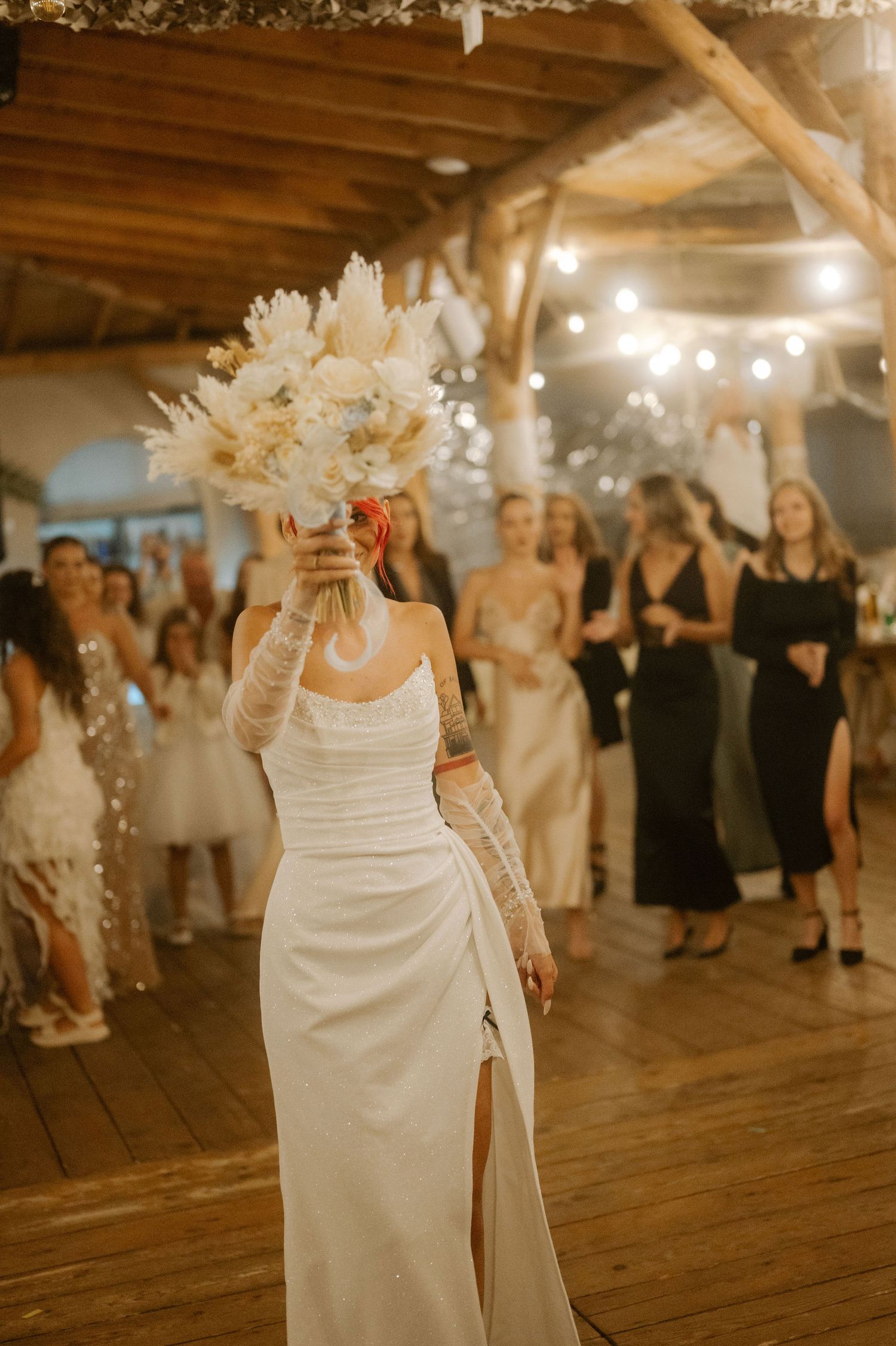 Wedding guests dance and celebrate on a rustic wooden dance floor illuminated by warm string lights overhead.