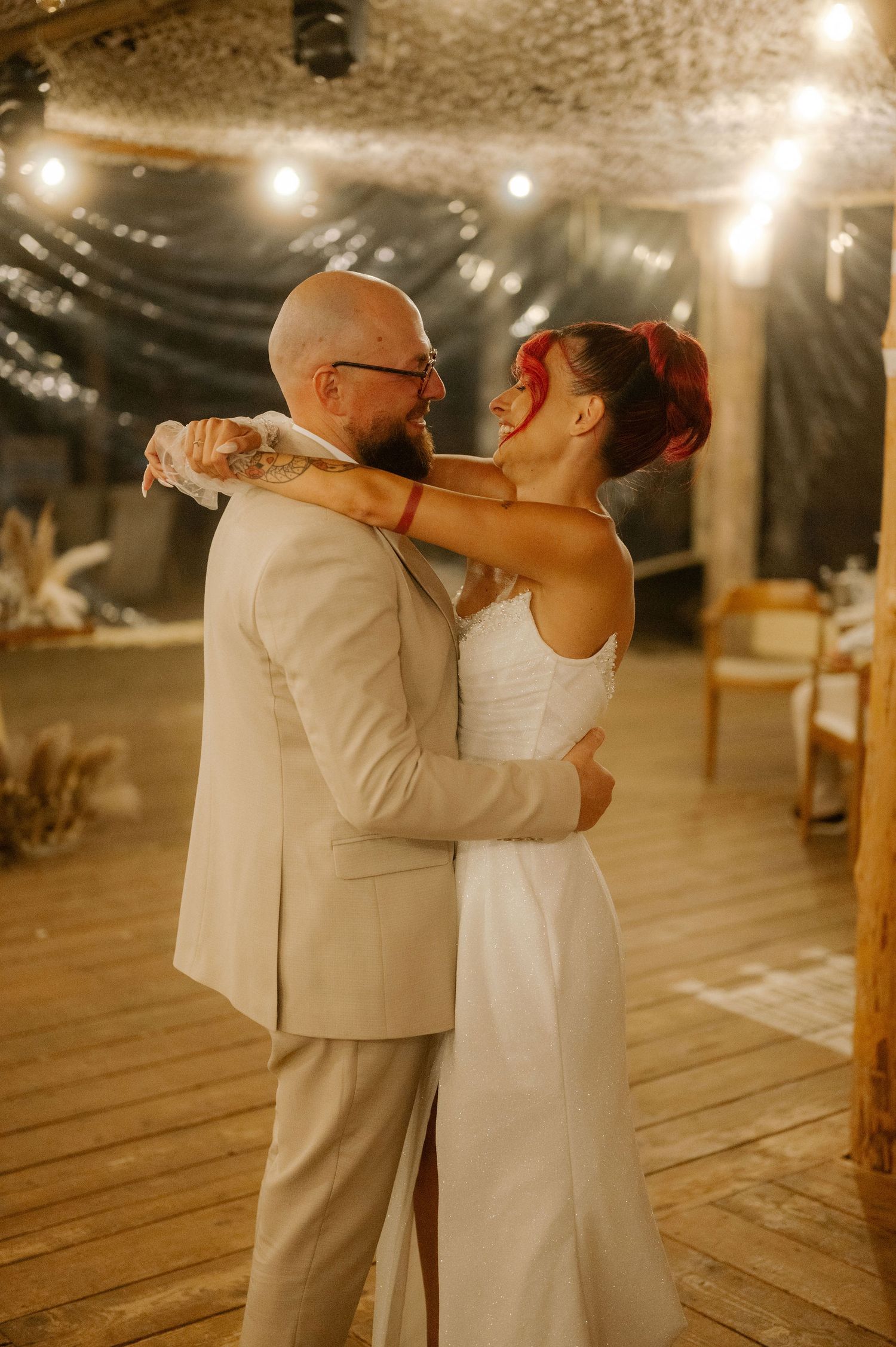 A couple shares a romantic dance moment under warm string lights in a rustic wooden venue.