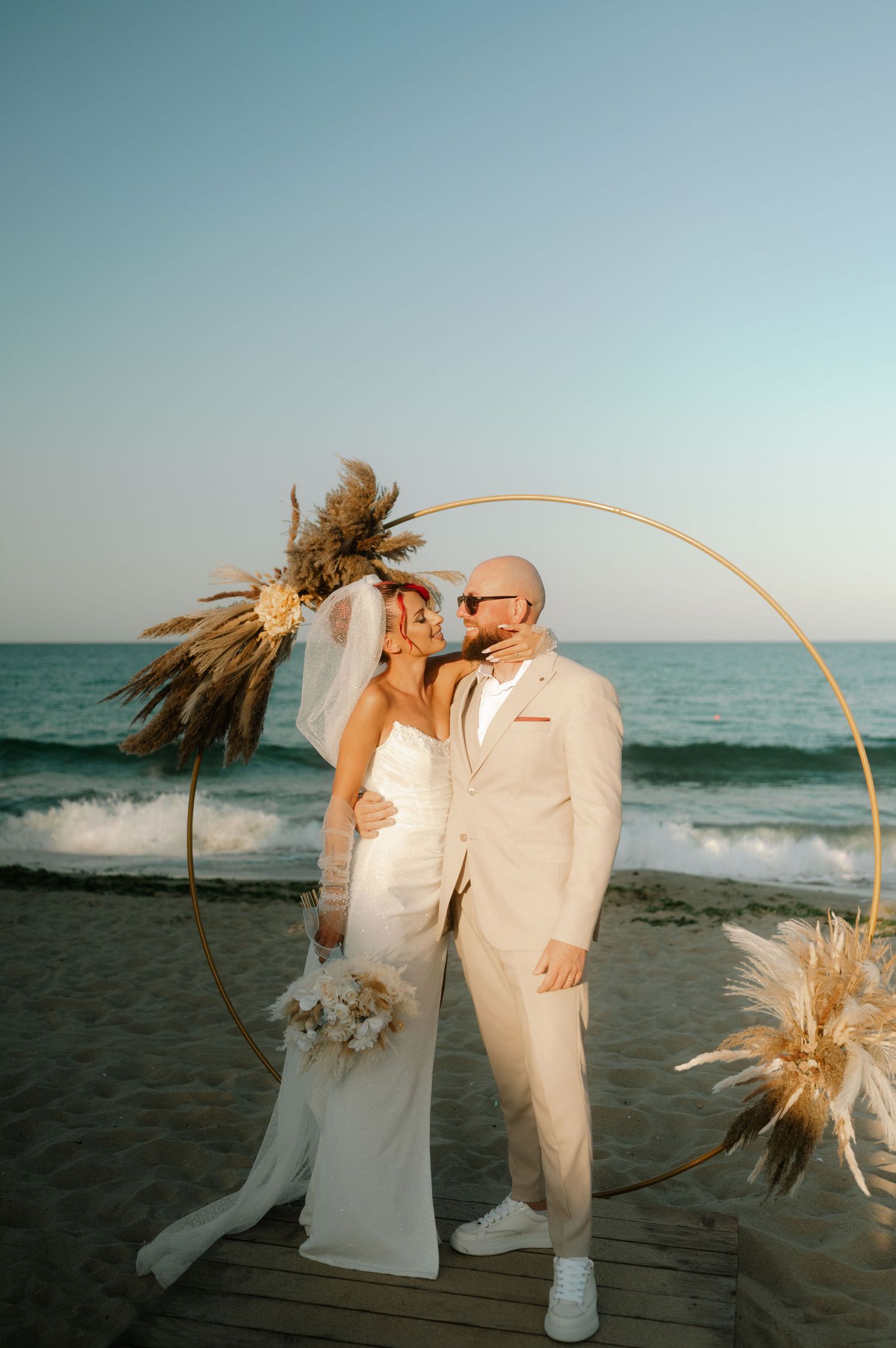 A beach wedding couple poses against a circular arch decorated with pampas grass as waves crash behind them at sunset.