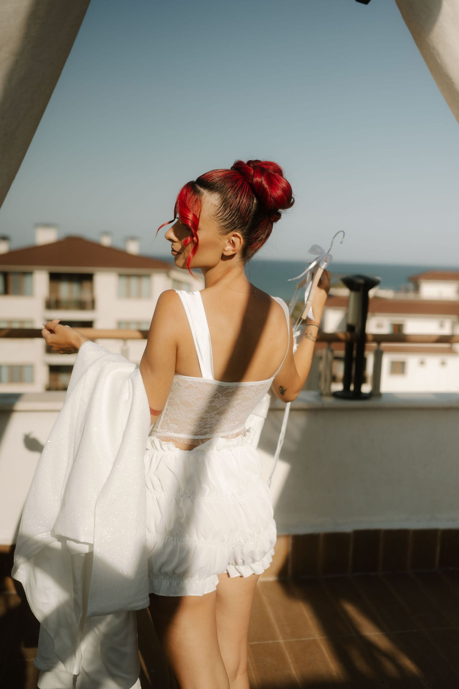 Person with bright red hair in white summer outfit stands on sunny balcony overlooking residential buildings.