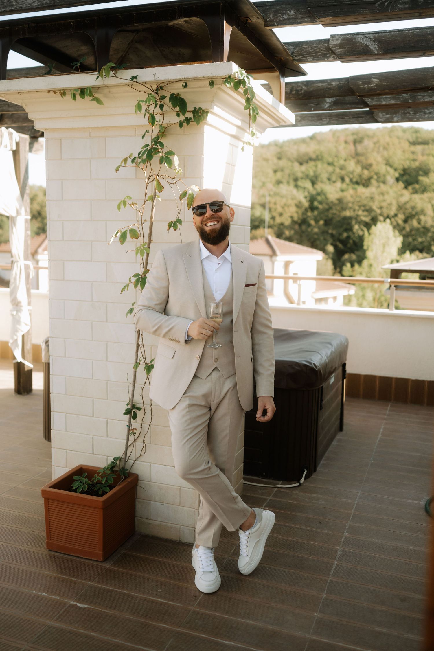 A person in a light beige suit and white sneakers leans against a white pillar on a terrace with climbing vines.