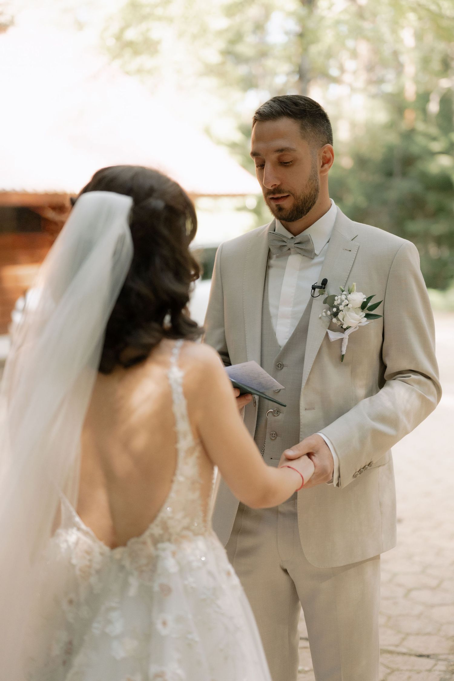 Groom in tan suit reads vows while holding bride's hands during outdoor wedding ceremony among tall pine trees.