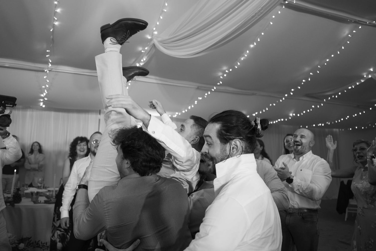 Guests lifting a man in the air during a lively wedding reception celebration in a decorated venue.