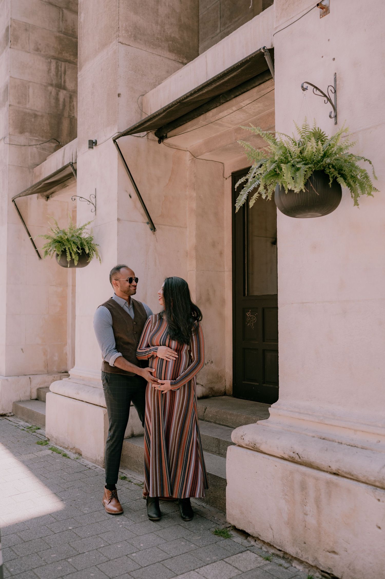 Couple posing outside a stone building with hanging fern plants, woman in striped maxi dress cradling baby bump.