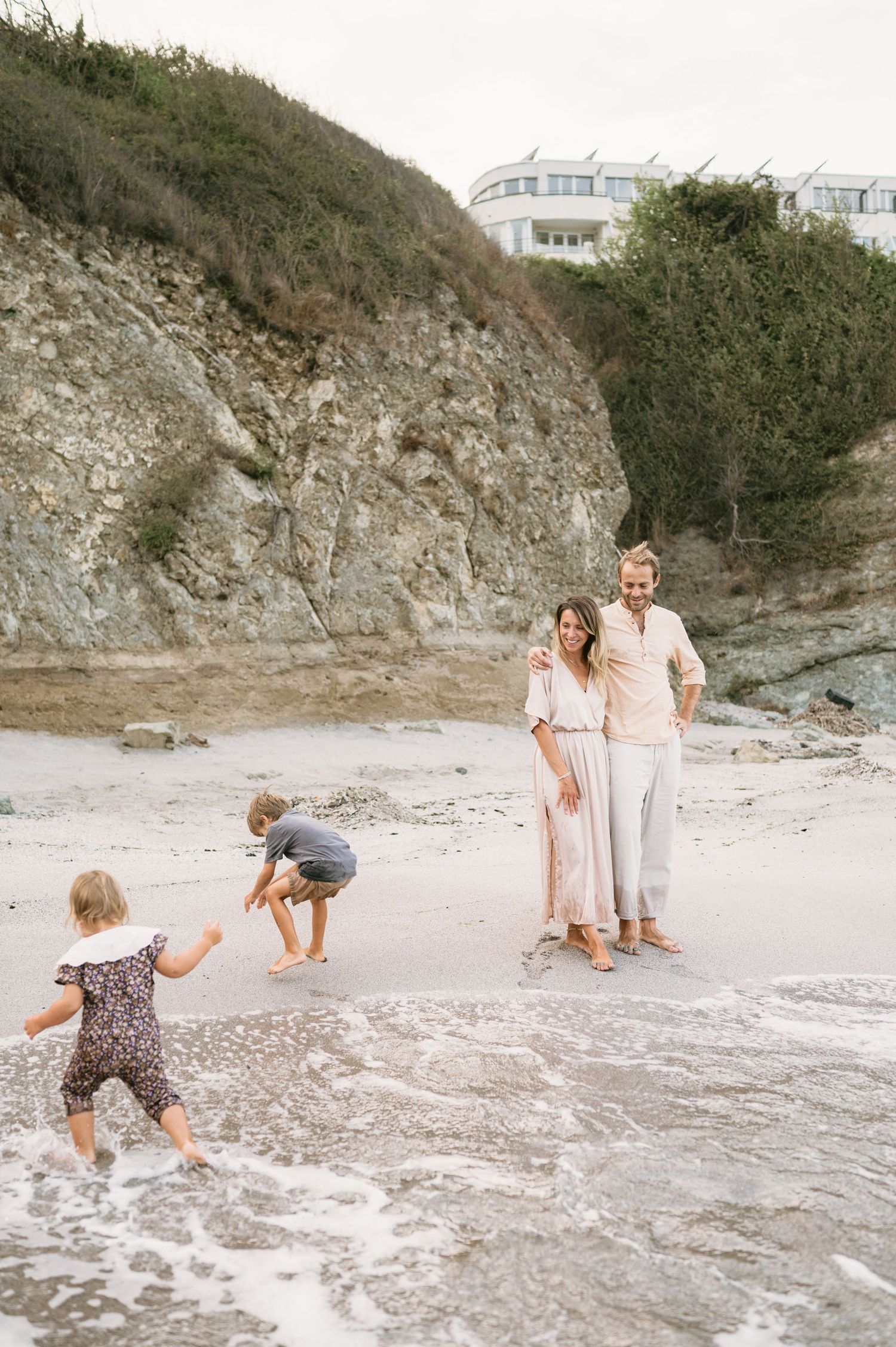 A family enjoys a beach day with two young children playing in the waves near rocky cliffs.