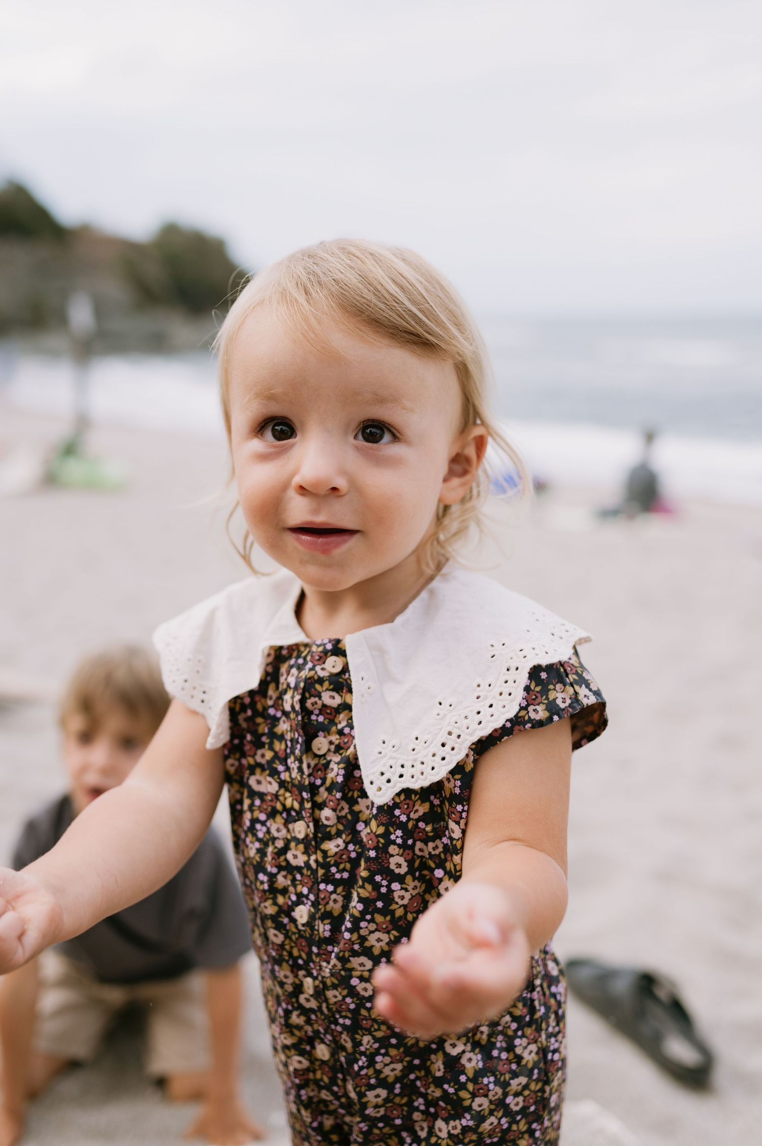 A toddler girl in a floral dress with white eyelet collar plays on a sandy beach on a cloudy day.