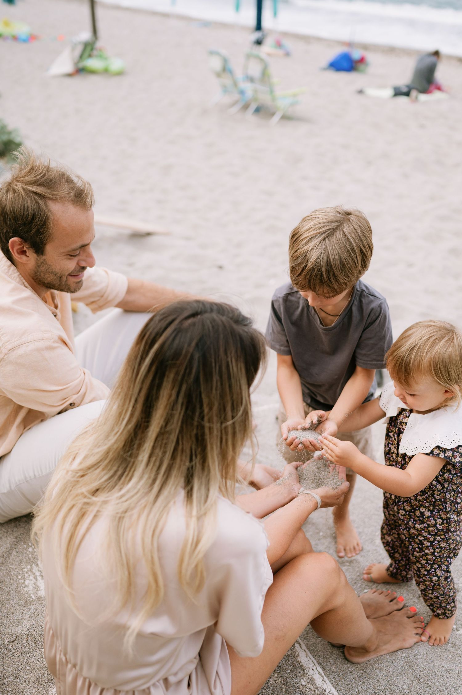 Family of four sitting on sandy beach steps, parents watching children play with sand on a sunny day.