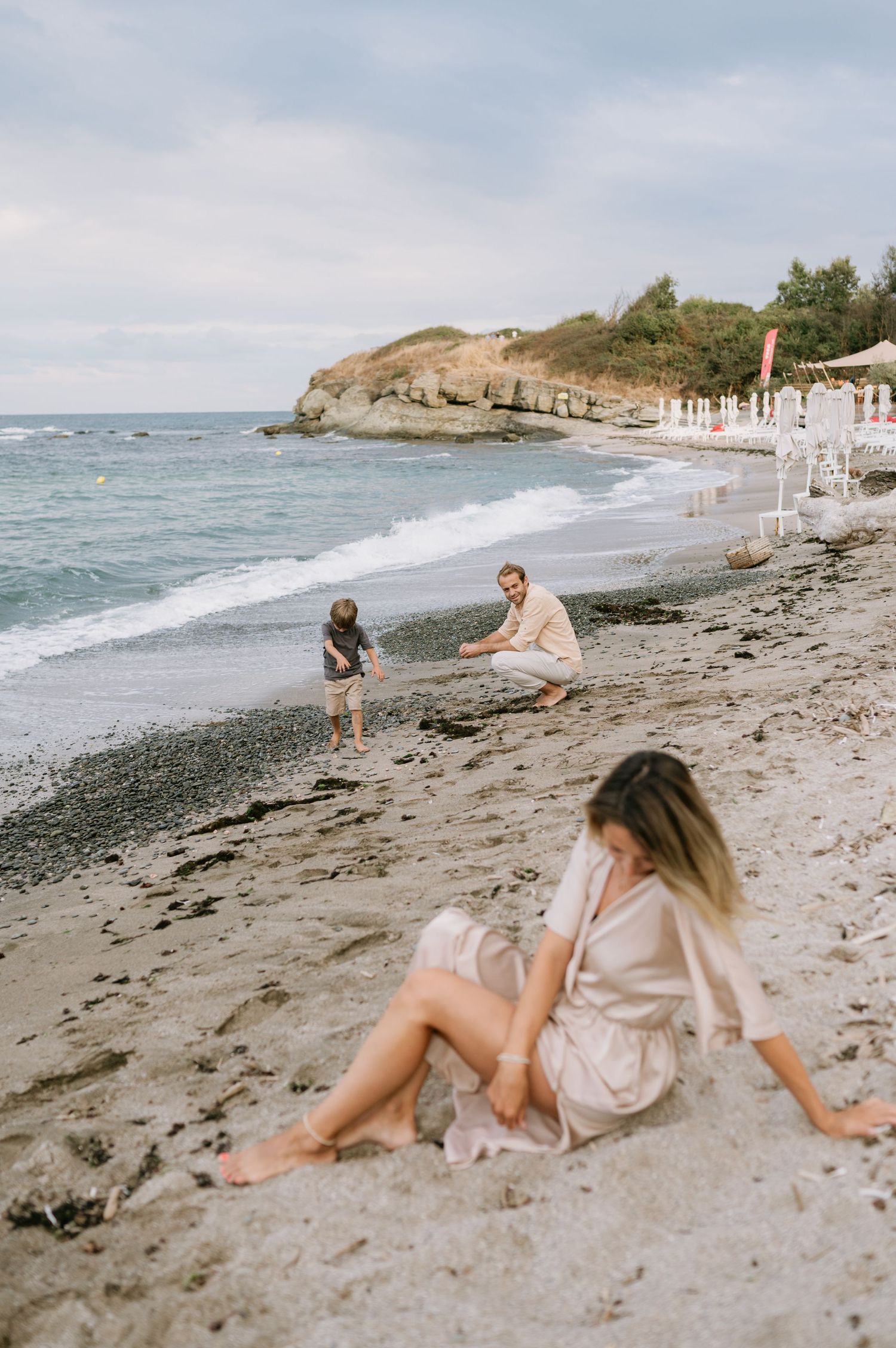 Family enjoying a beach day, woman sitting on sandy shore while man plays with young child near ocean waves.