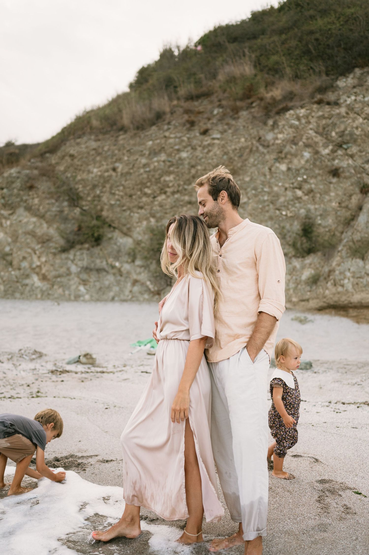 A couple shares a romantic moment on a rocky beach while two young children play in the sand nearby.