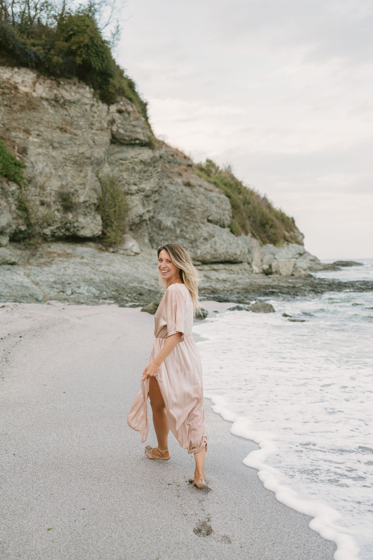 Woman in pink maxi dress walking on sandy beach with rocky cliffs and ocean waves in background.