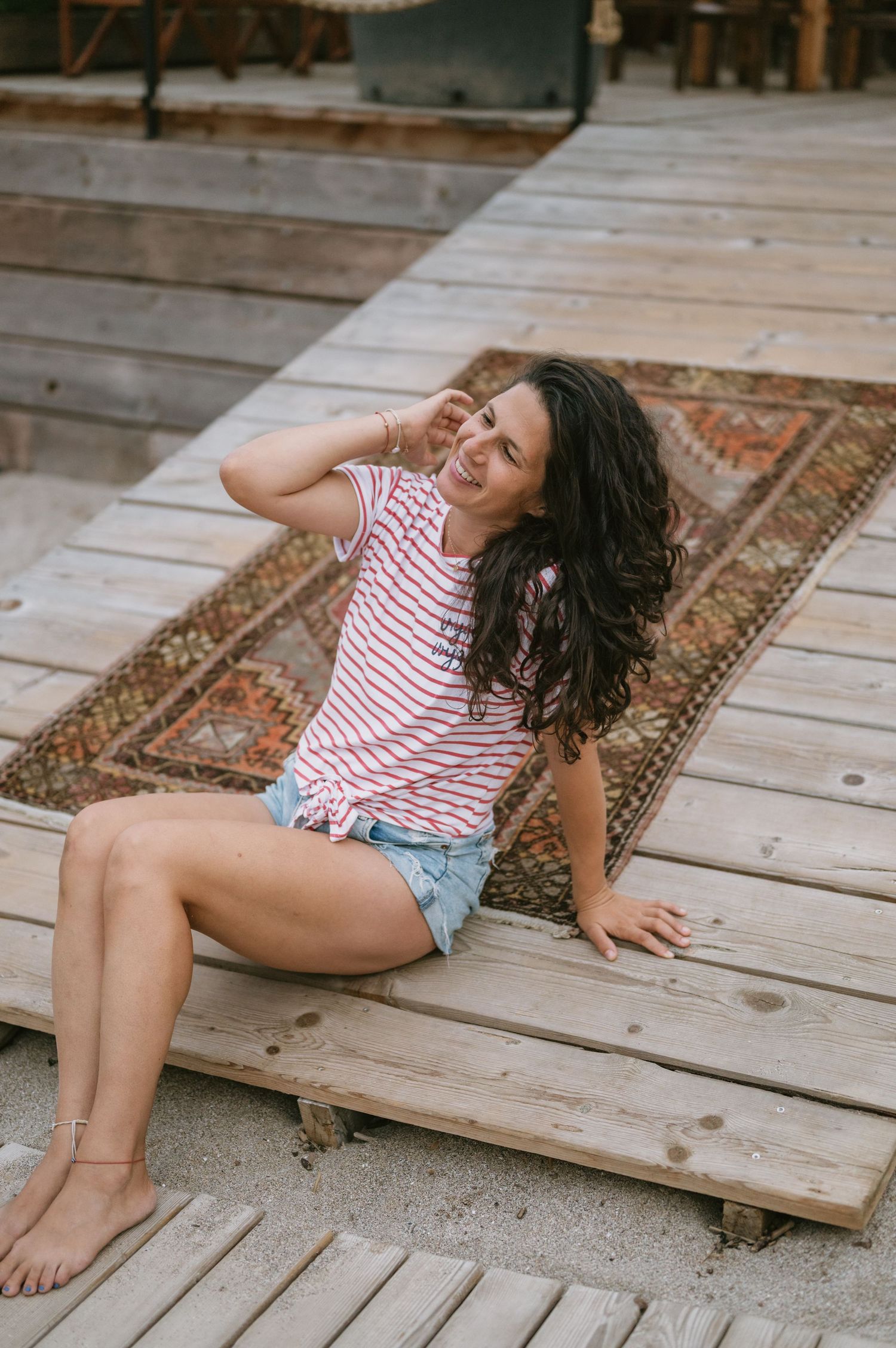 Smiling brunette woman sitting on wooden deck wearing striped tank top and denim shorts with a vintage rug behind her.