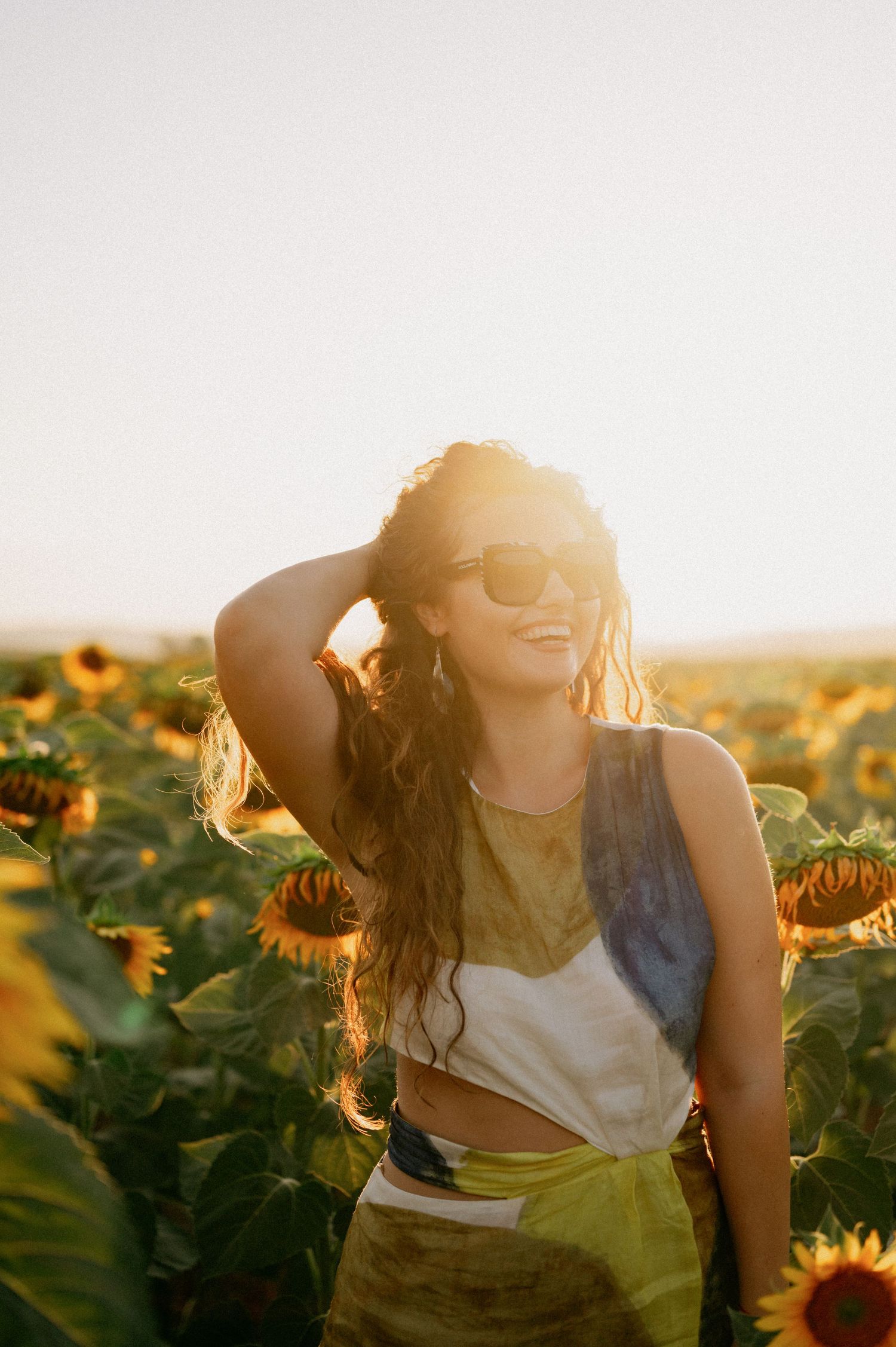 Woman smiling in sunflower field at golden hour, wearing sunglasses and tie-dye crop top, hand in curly hair.