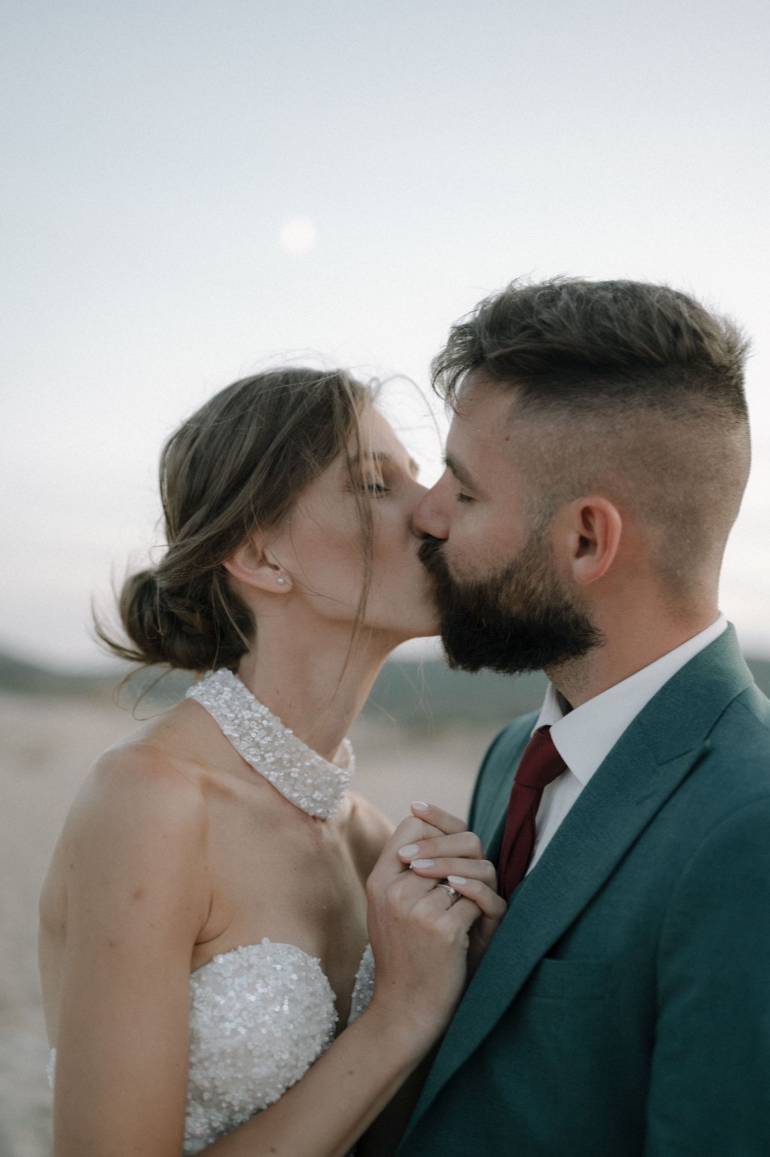 A bride and groom share a romantic kiss on a beach, she in a beaded halter gown, he in a teal suit with red tie.