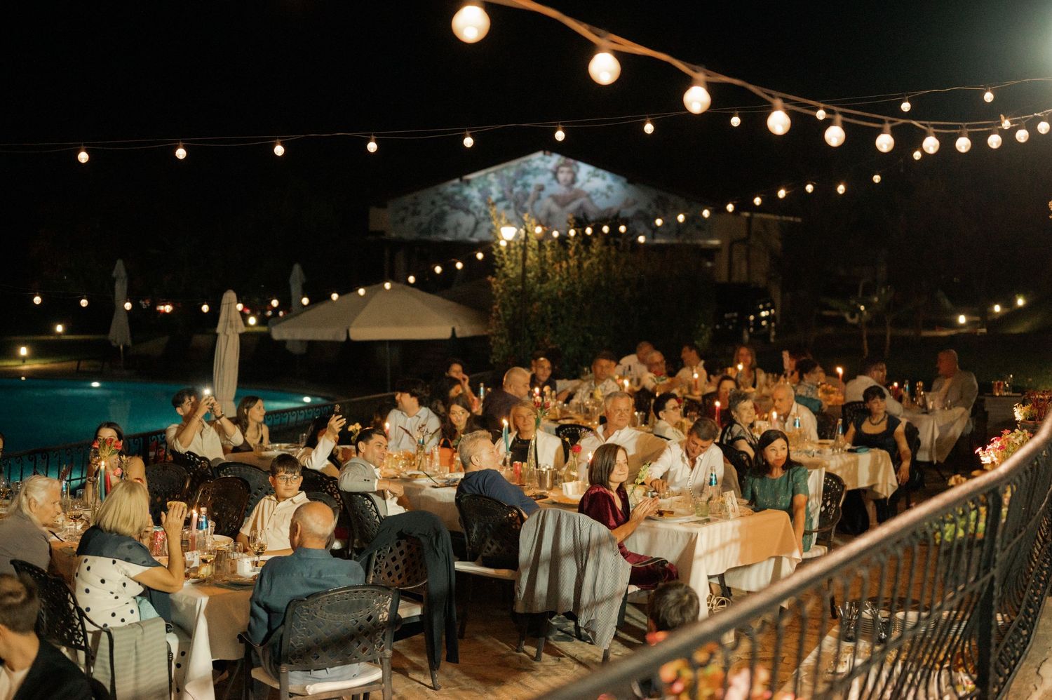 Outdoor evening reception with string lights illuminating guests dining at tables on a patio.