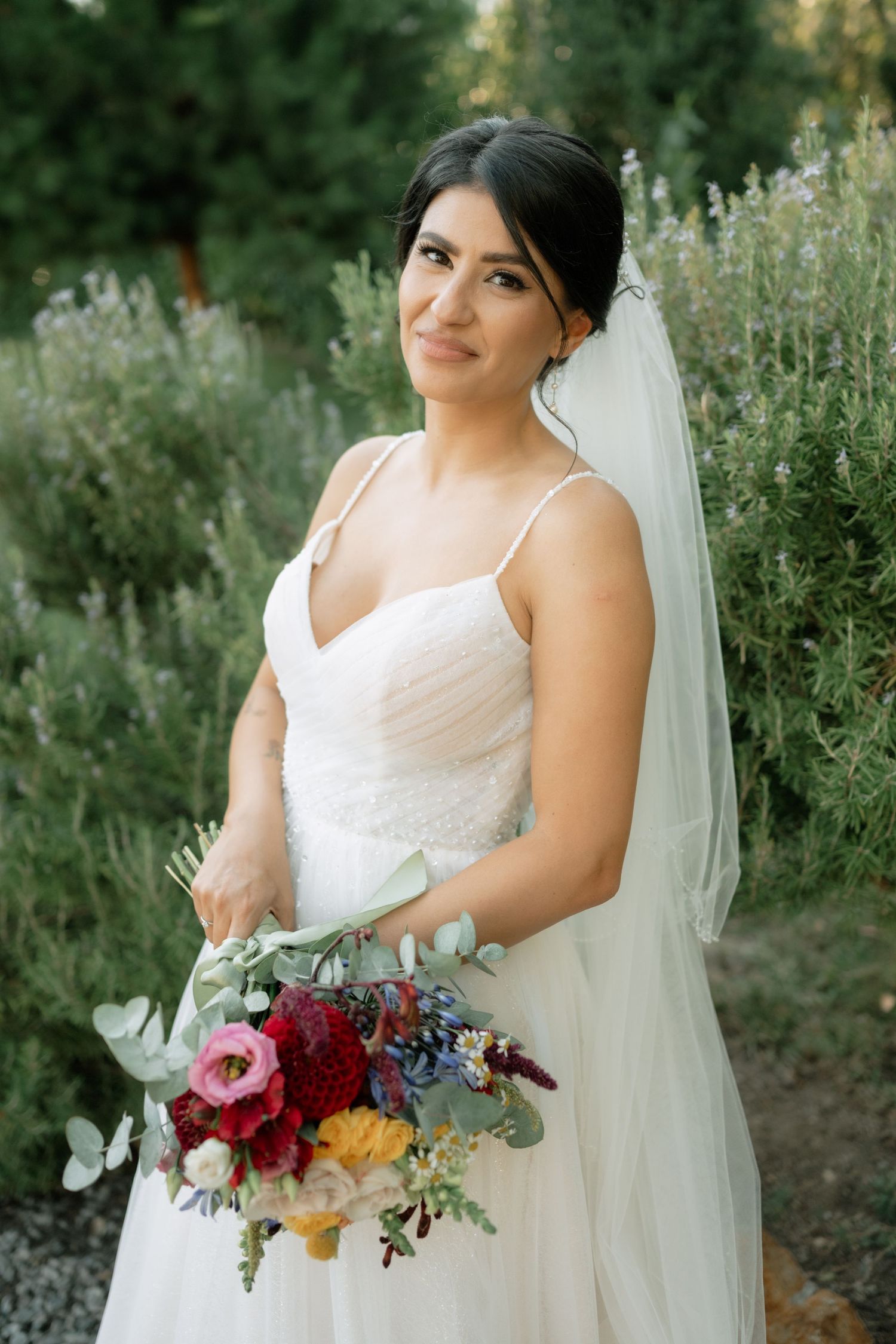 A bride in a white wedding dress holds a vibrant bouquet of red and burgundy flowers against a natural outdoor backdrop.
