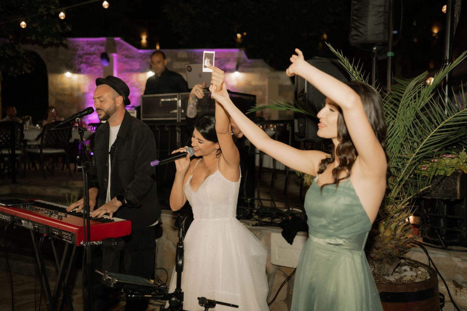 People dancing and celebrating at a nighttime party with string lights and palm decor in the background.