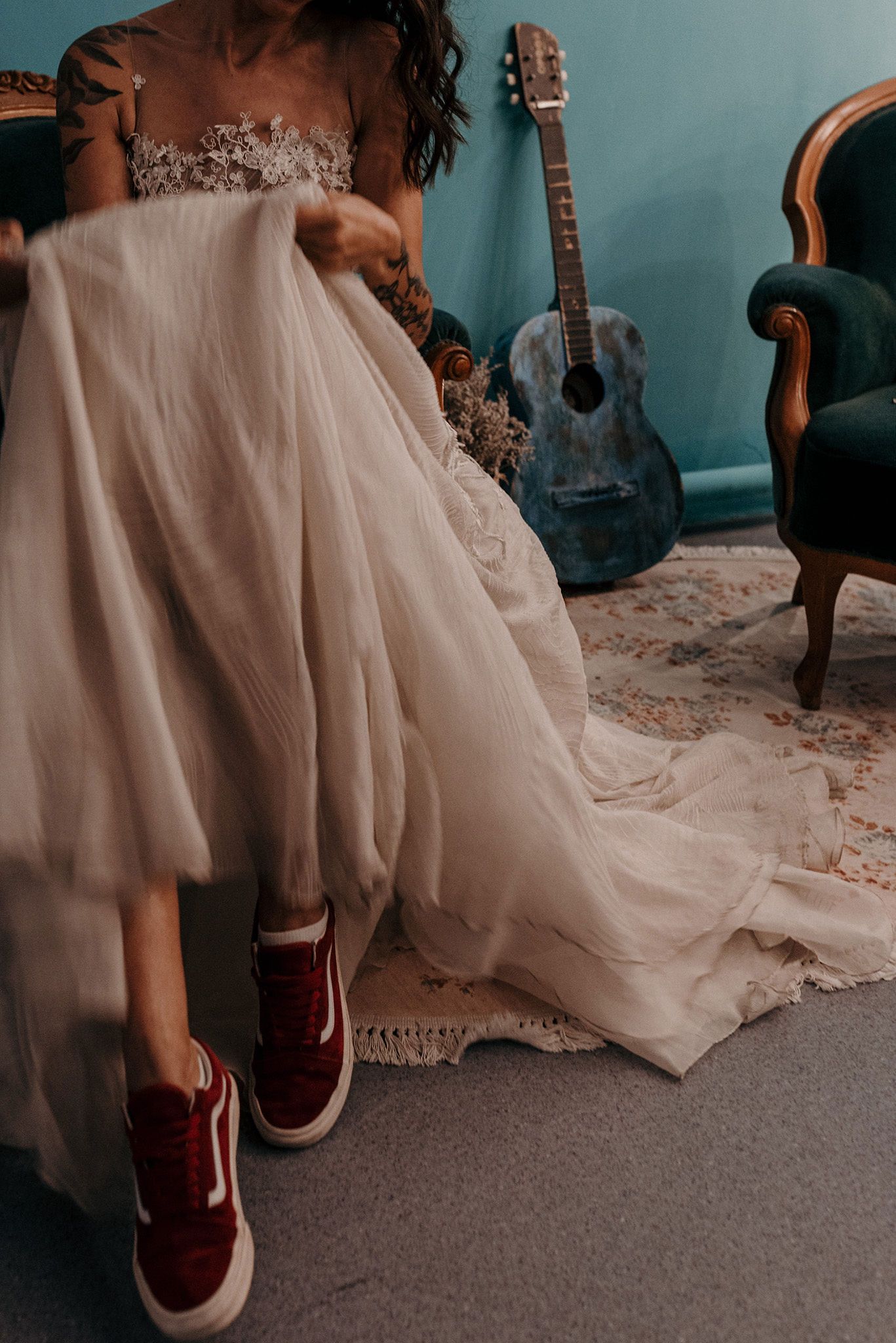 Wedding dress with burgundy sneakers and acoustic guitar against teal wall and vintage furniture.