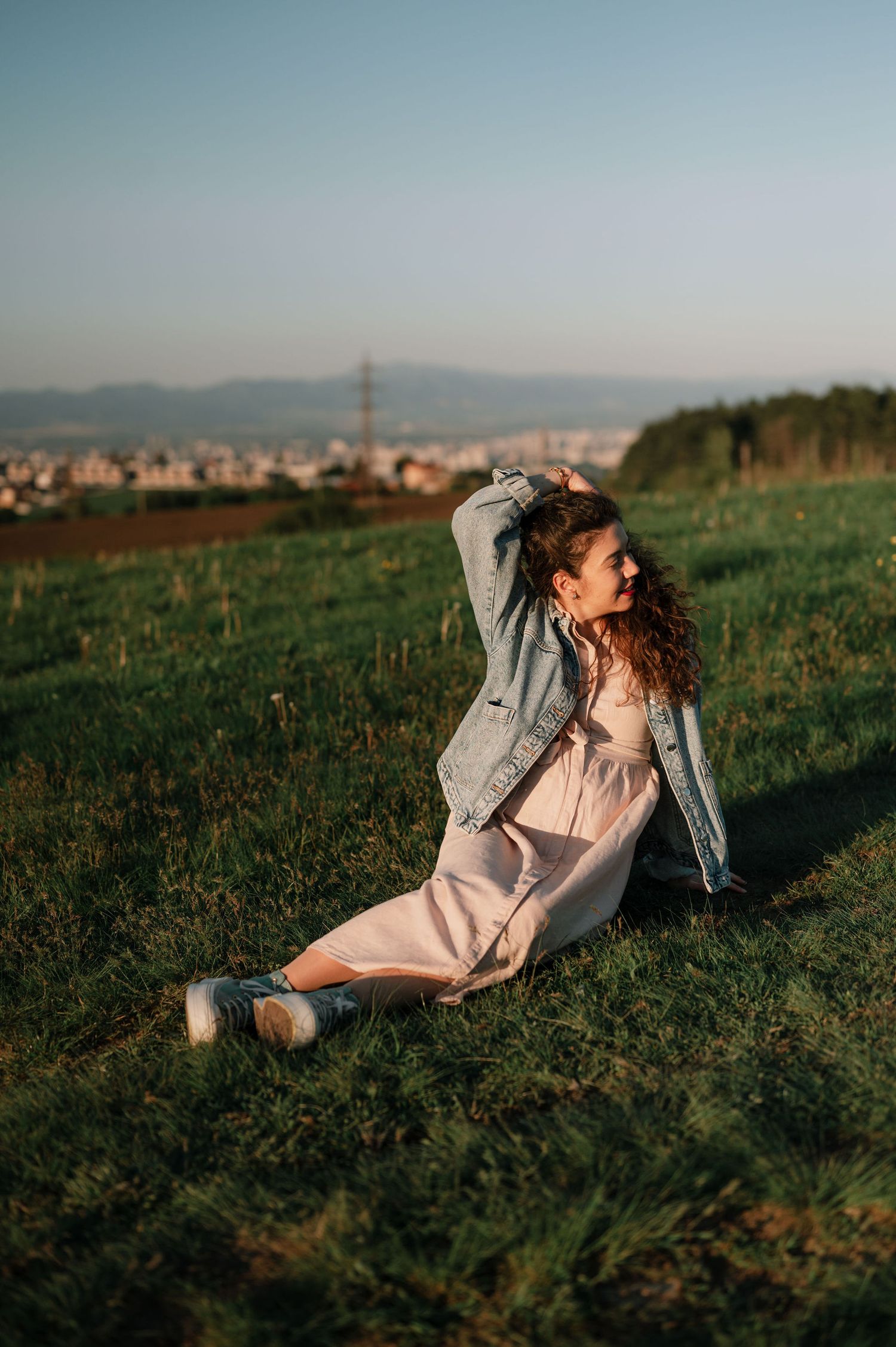 Person in white dress and denim jacket lounges in grassy field during sunset with city skyline in background.
