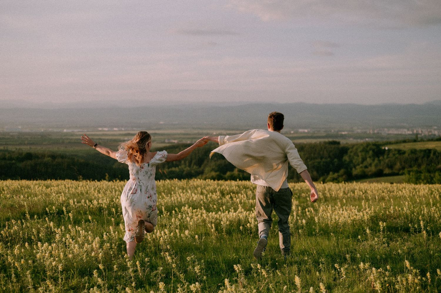 Two people run joyfully through a wildflower field with arms outstretched at sunset.