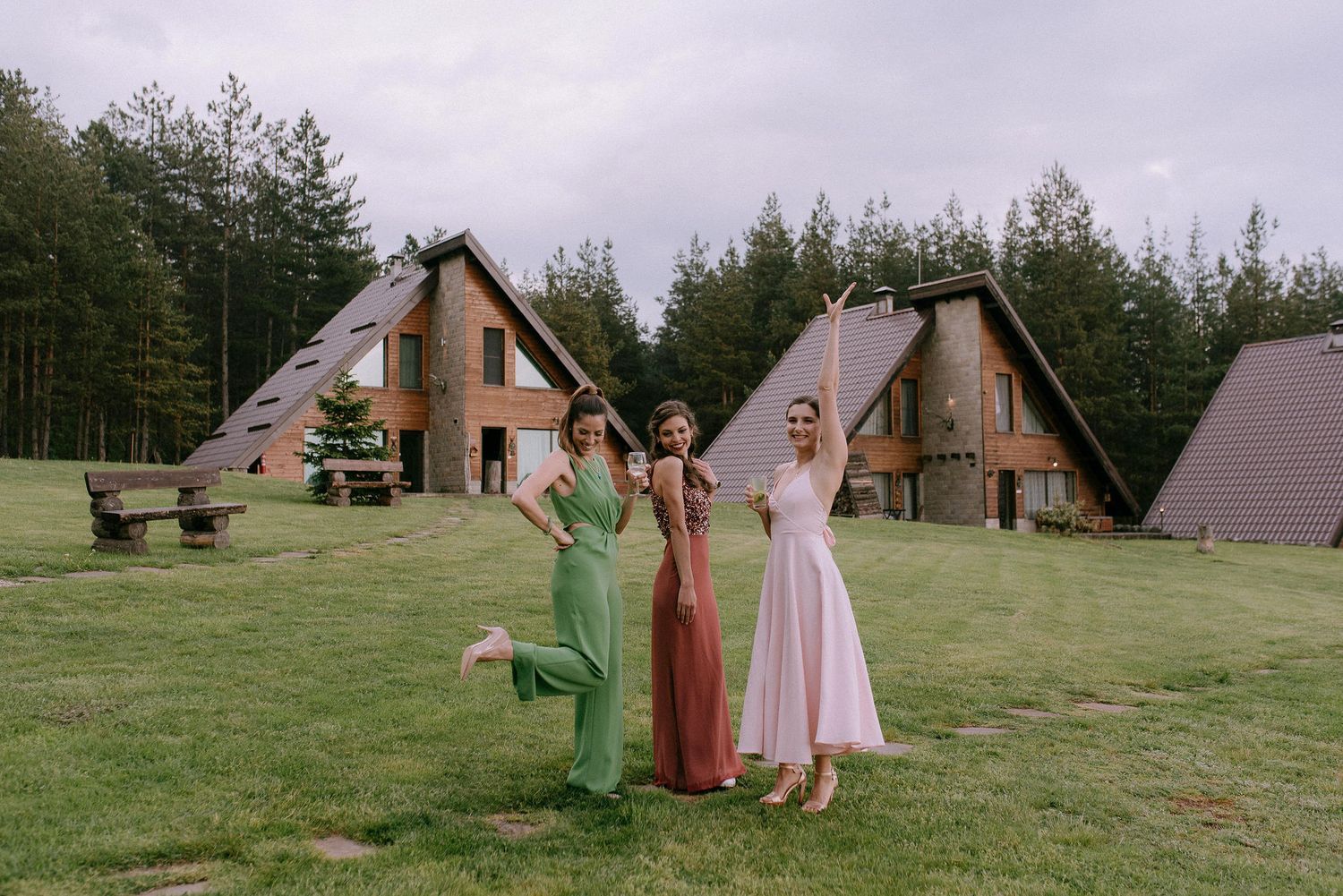 Three people in formal attire pose joyfully in front of A-frame cabins in a grassy forest setting.