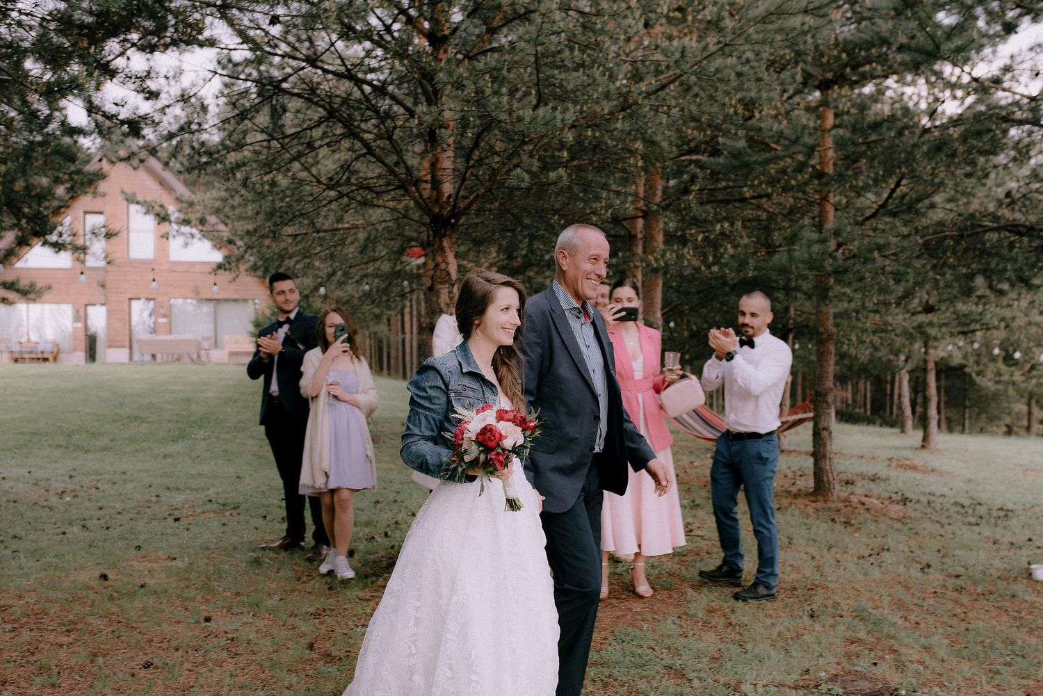 Outdoor wedding celebration on a grassy field with pine forest and cabin backdrop.