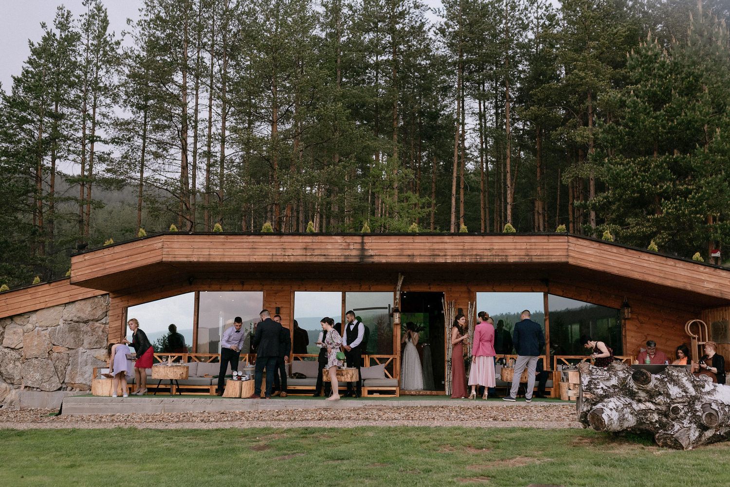 Modern wooden and stone wedding venue nestled among pine trees with guests gathered outside.