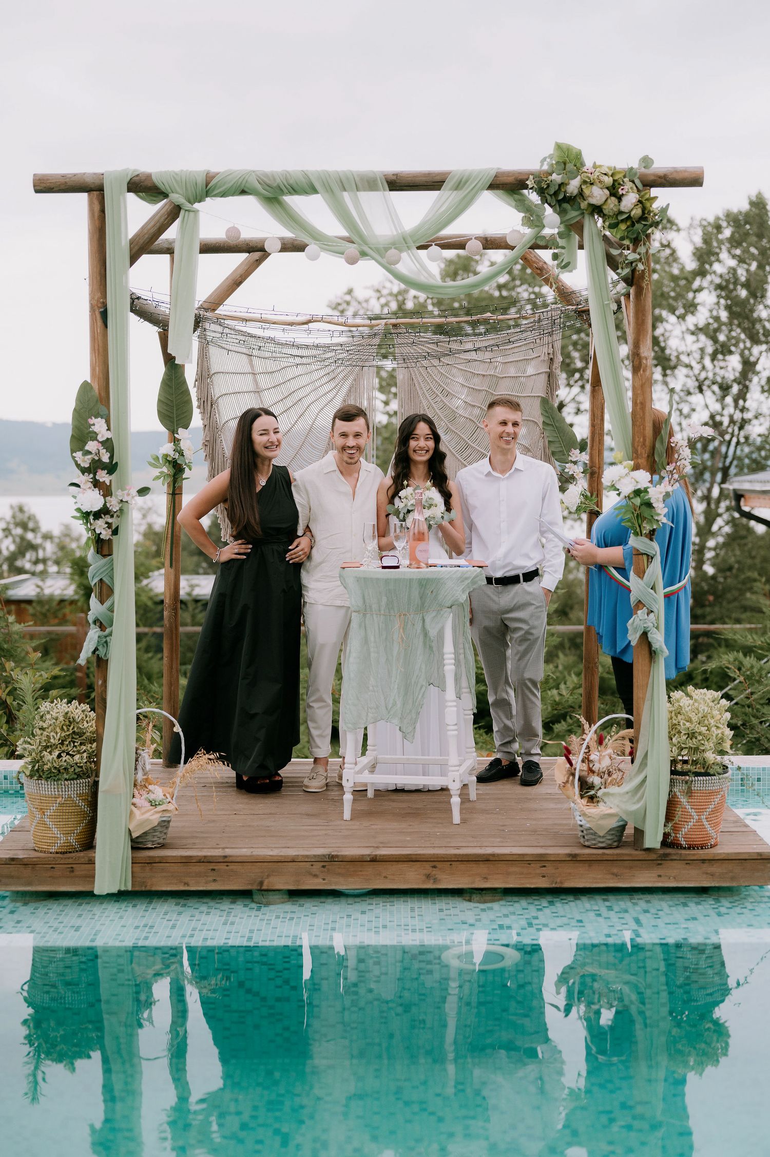 Wedding ceremony taking place under a decorated wooden pergola by a turquoise pool.