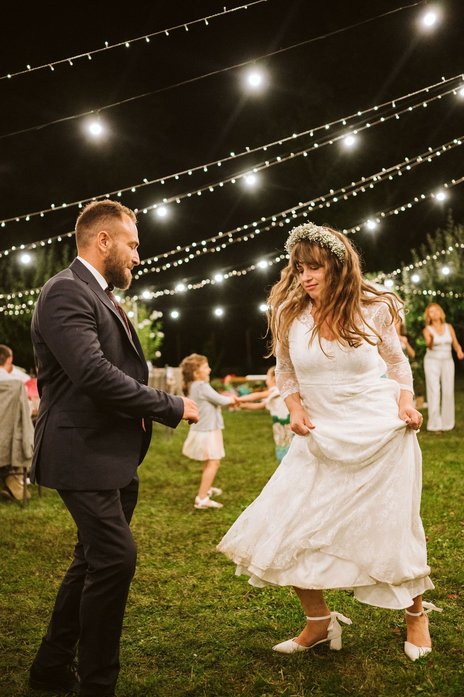 A couple dances joyfully under twinkling string lights at an outdoor evening wedding reception.