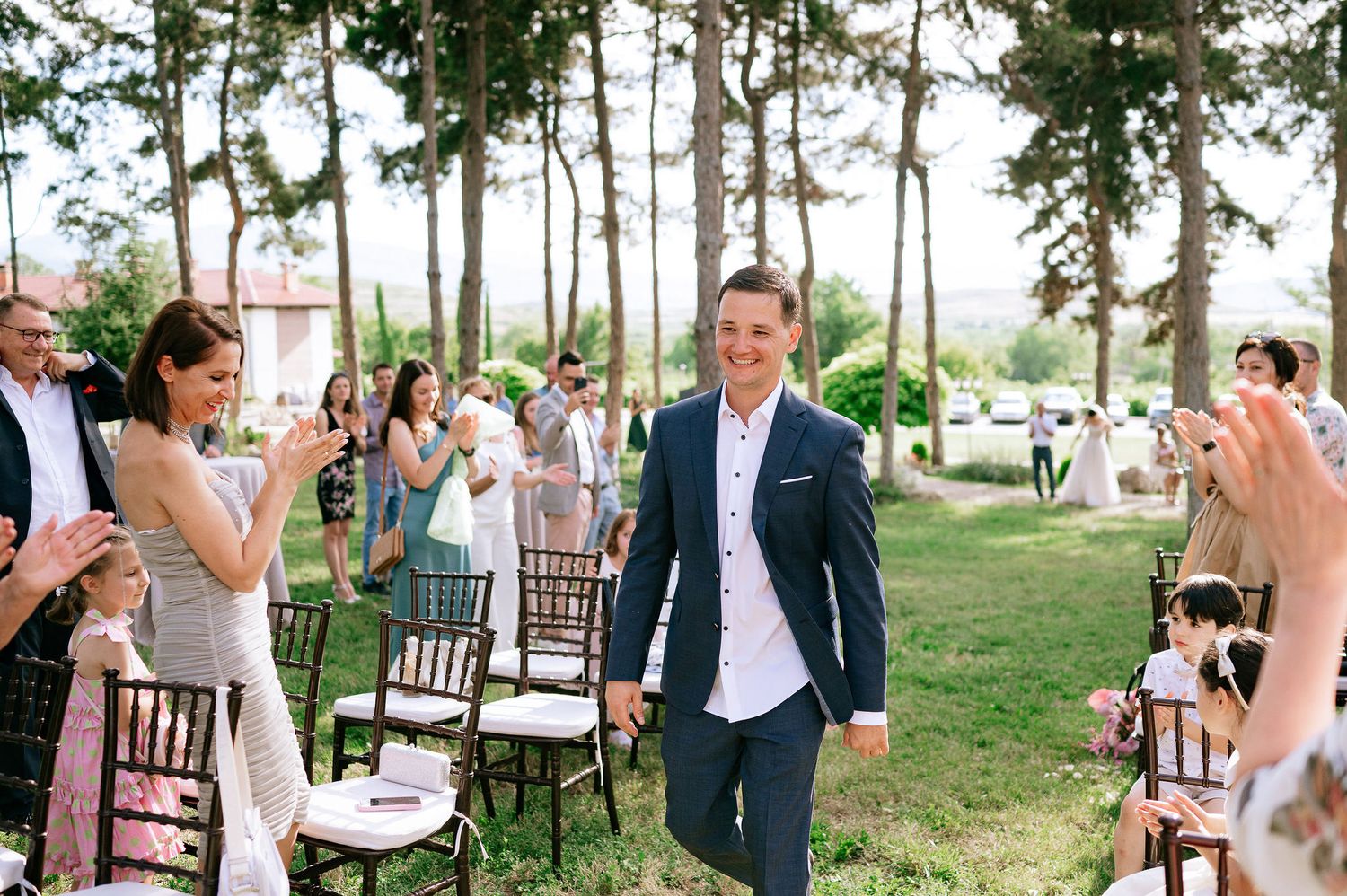 Wedding ceremony processional in progress with guests seated along aisle beneath tall trees.