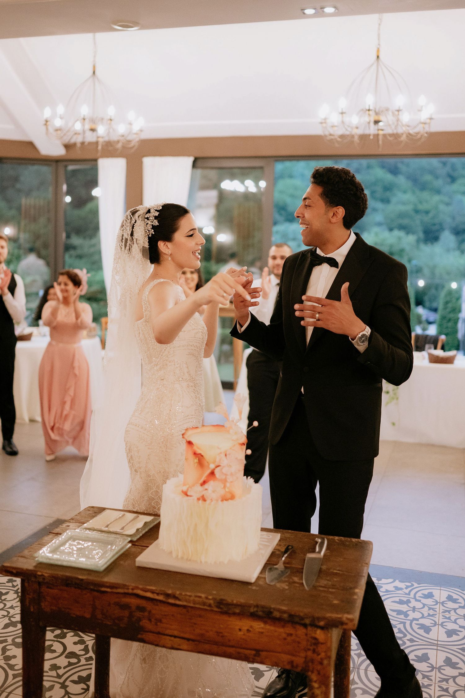 Newlyweds share a cake-cutting moment at their reception by a rustic wooden table.
